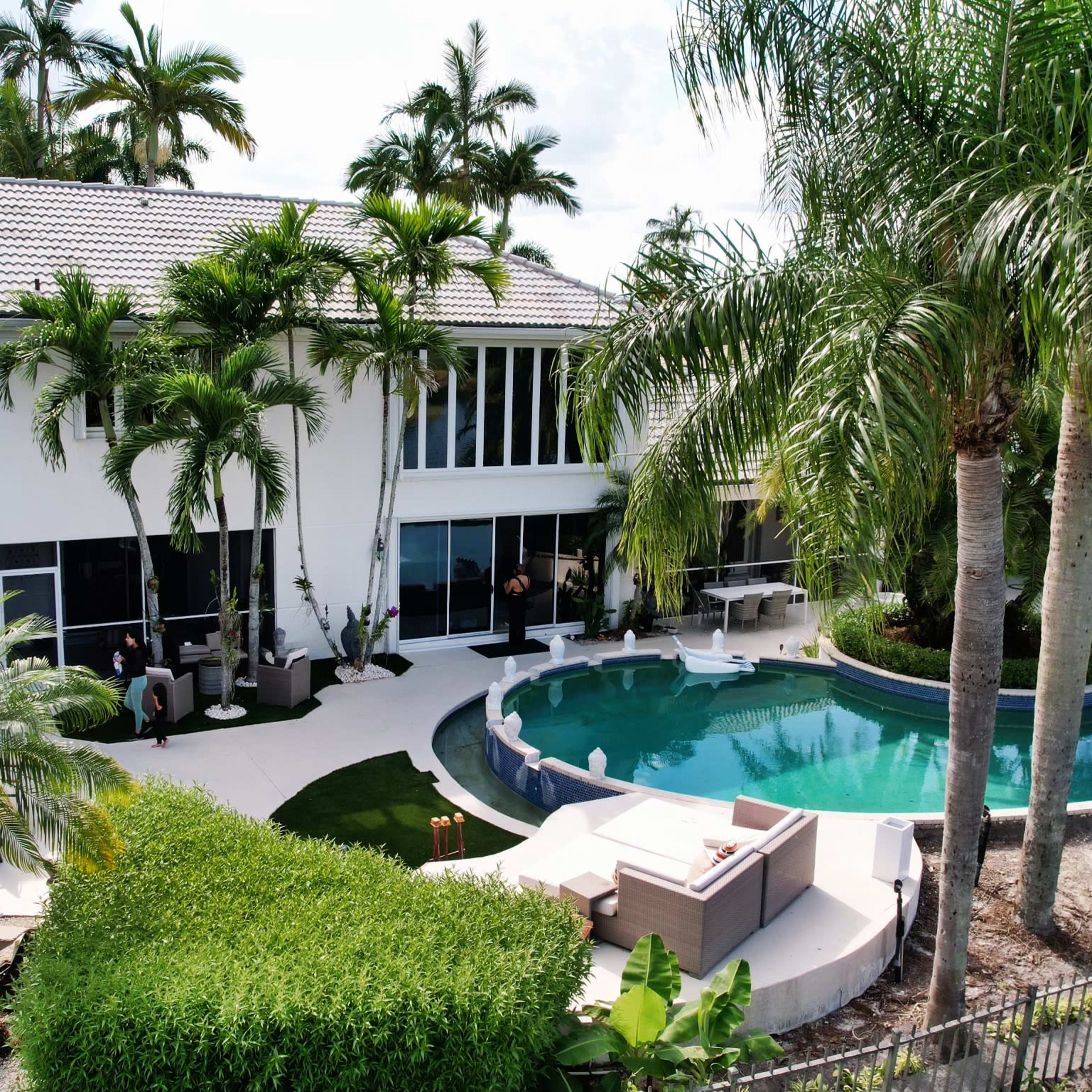 A modern two-story house surrounded by palm trees, a swimming pool, and landscaped gardens.