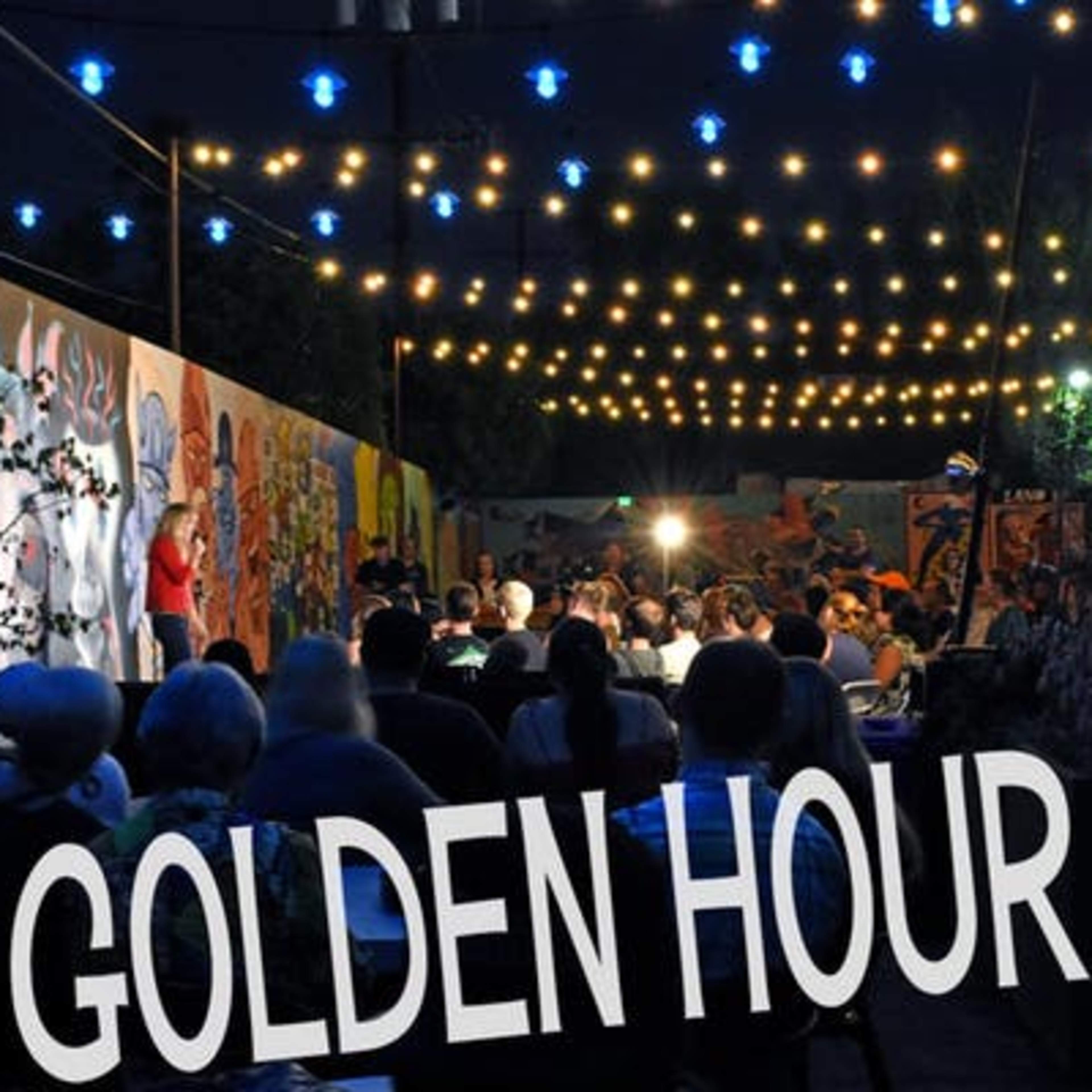 A lively outdoor gathering is illuminated by string lights, with a mural backdrop and an audience seated at tables.