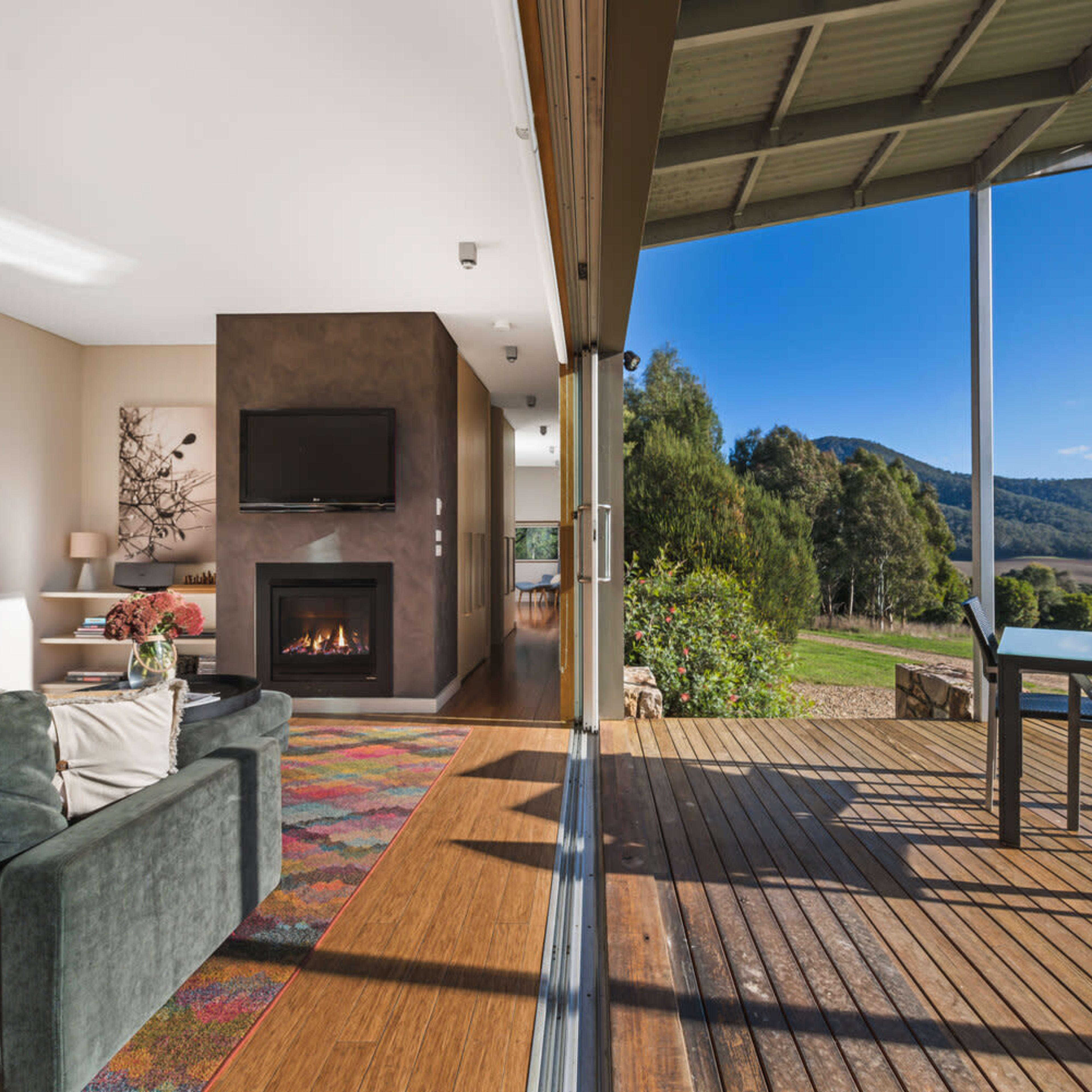 A modern living room with a fireplace and large windows opens to a wooden deck overlooking a mountainous landscape.