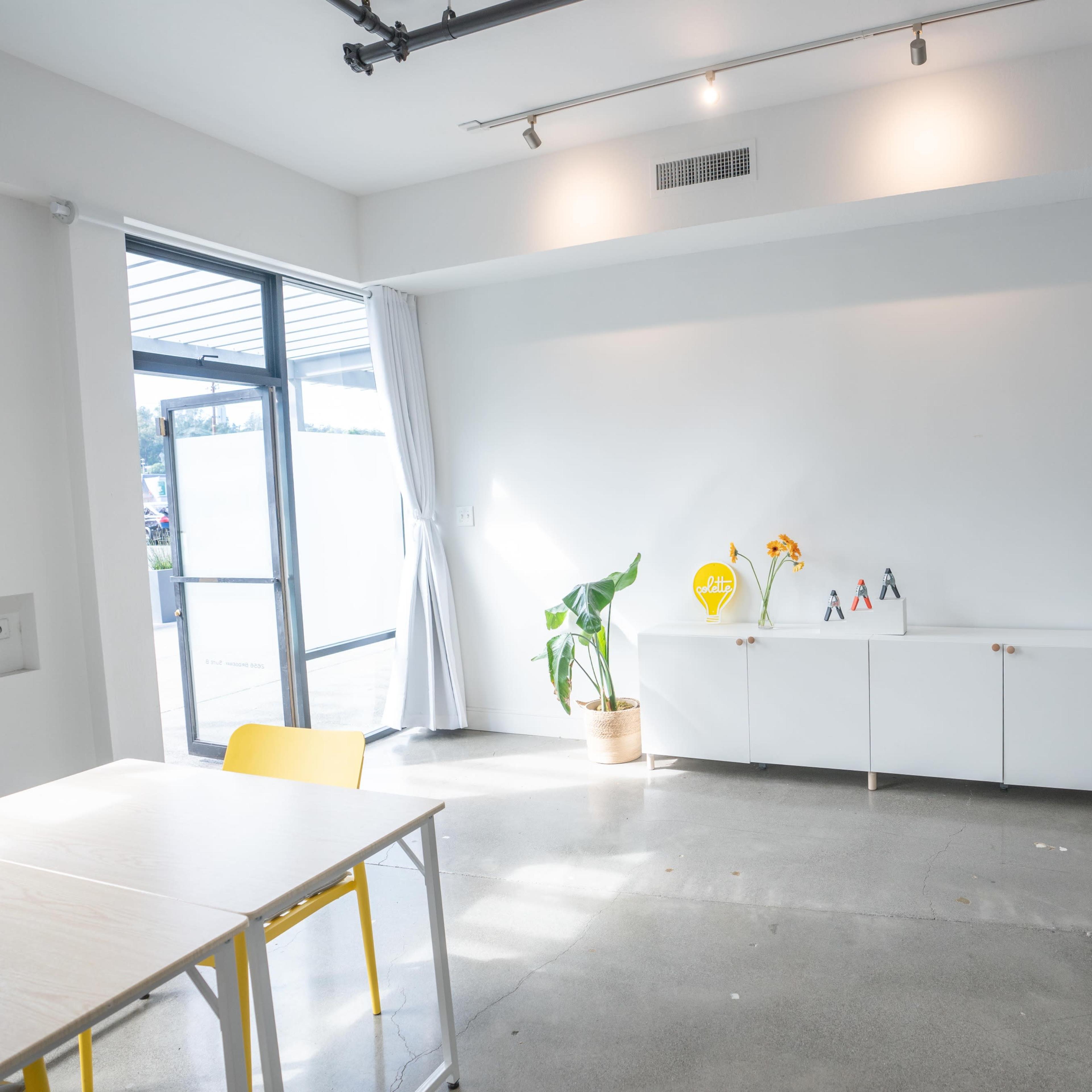 A bright, minimalist interior with a table and chairs, a potted plant, and a cabinet displaying small figures.