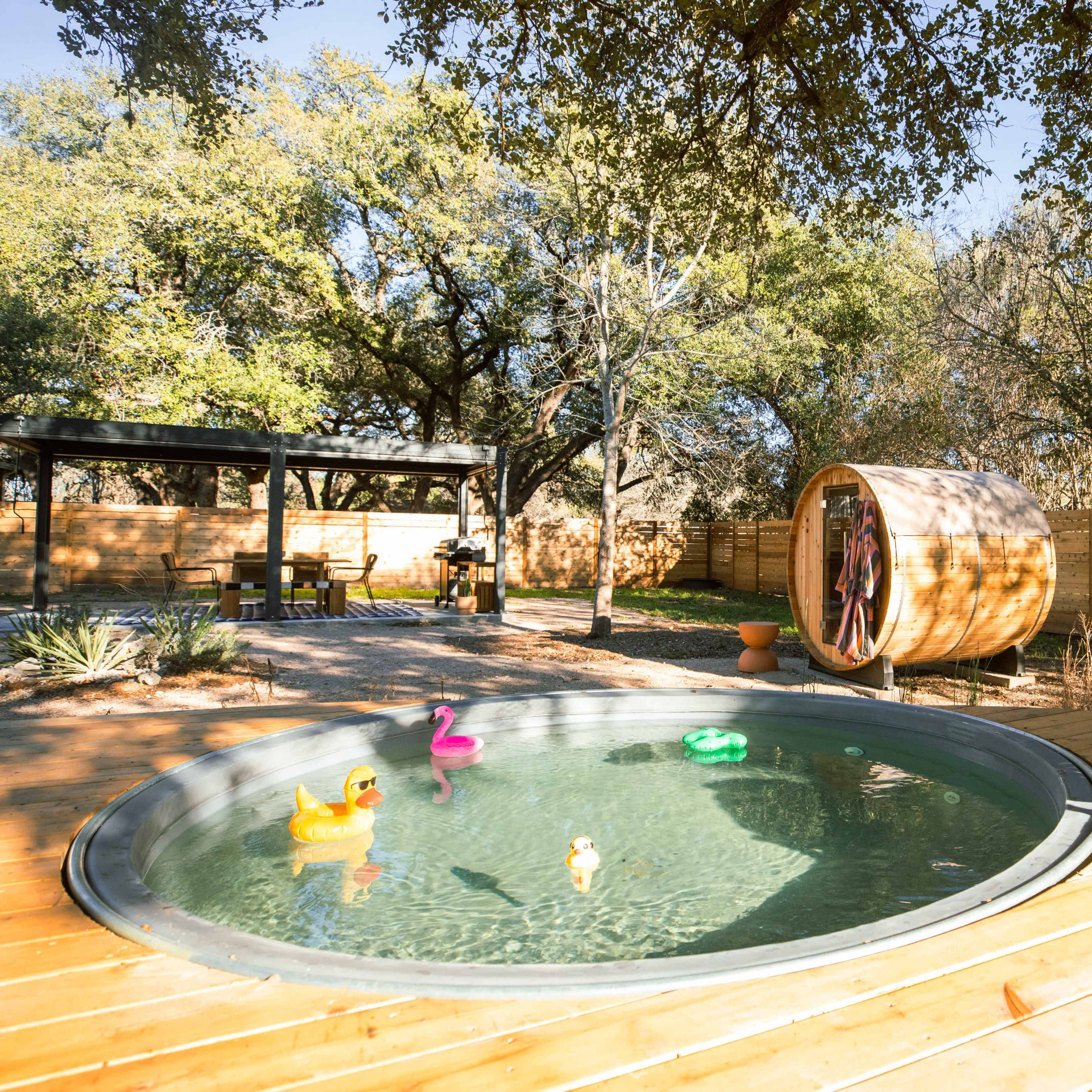 The scene features a circular hot tub surrounded by wooden decking, with colorful rubber ducks floating in the water and a large wooden barrel nearby.