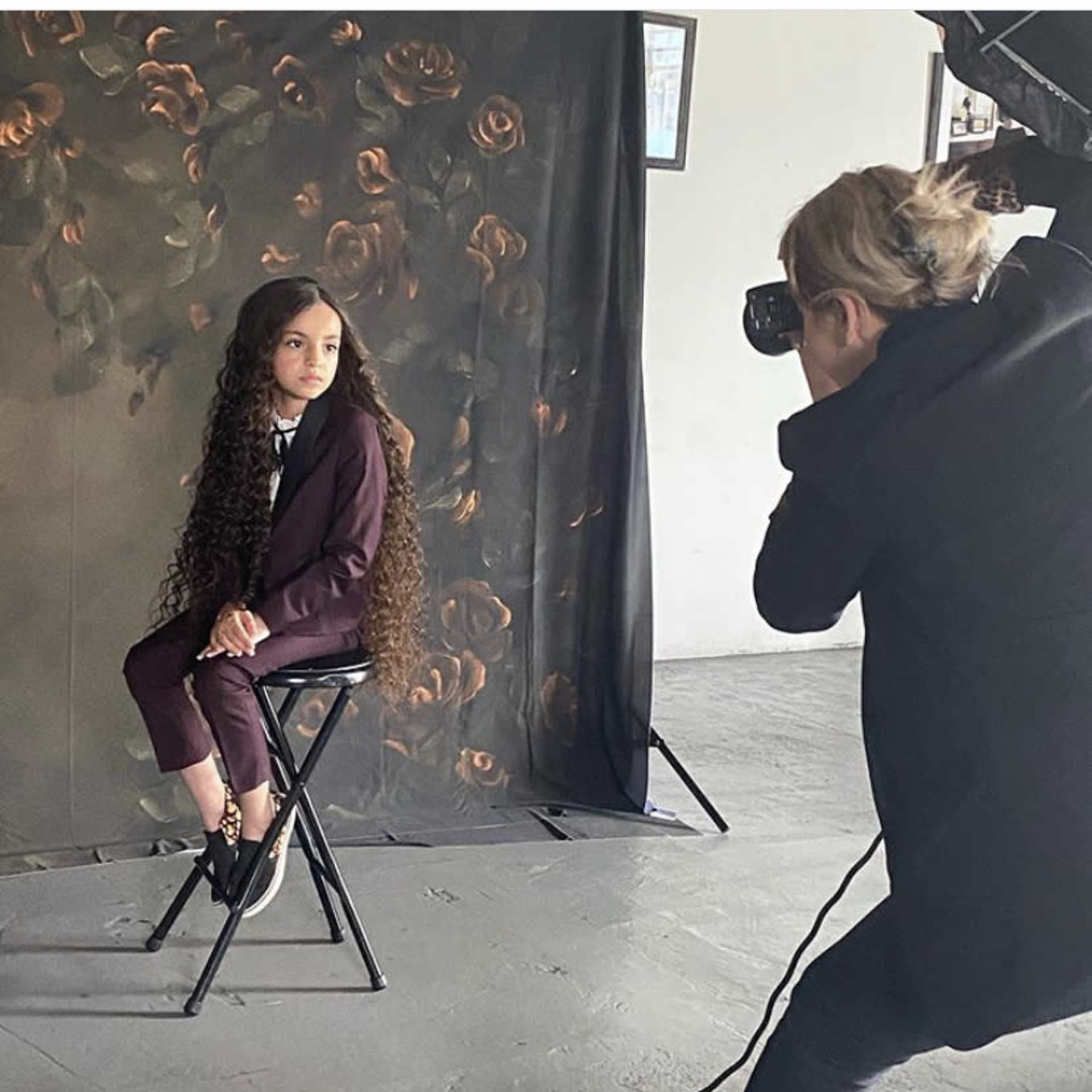 A child dressed in a suit sits on a stool while a photographer takes her picture in front of a floral backdrop.