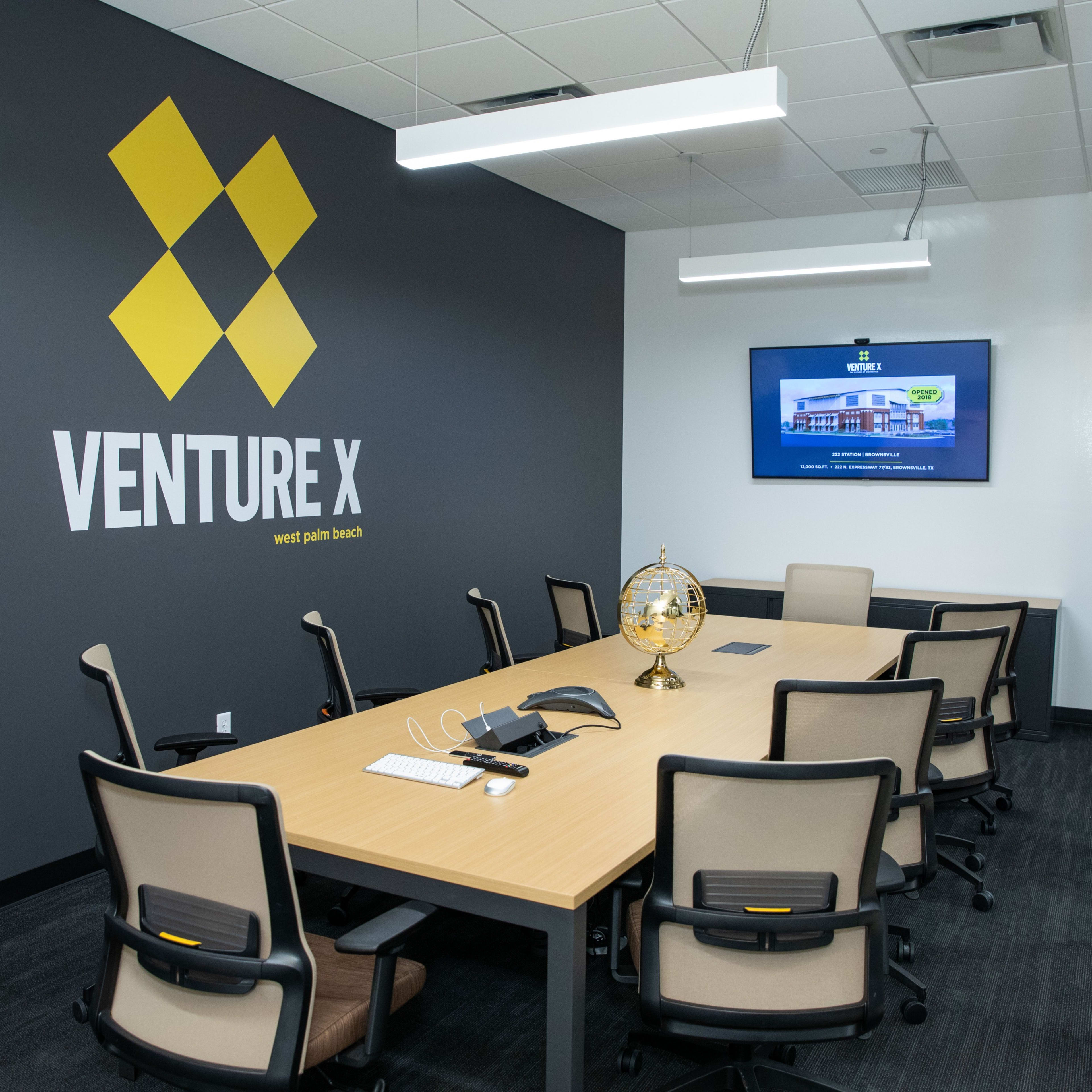 A modern conference room with a large table, several chairs, a wall-mounted screen, and a wall featuring the "Venture X" logo.