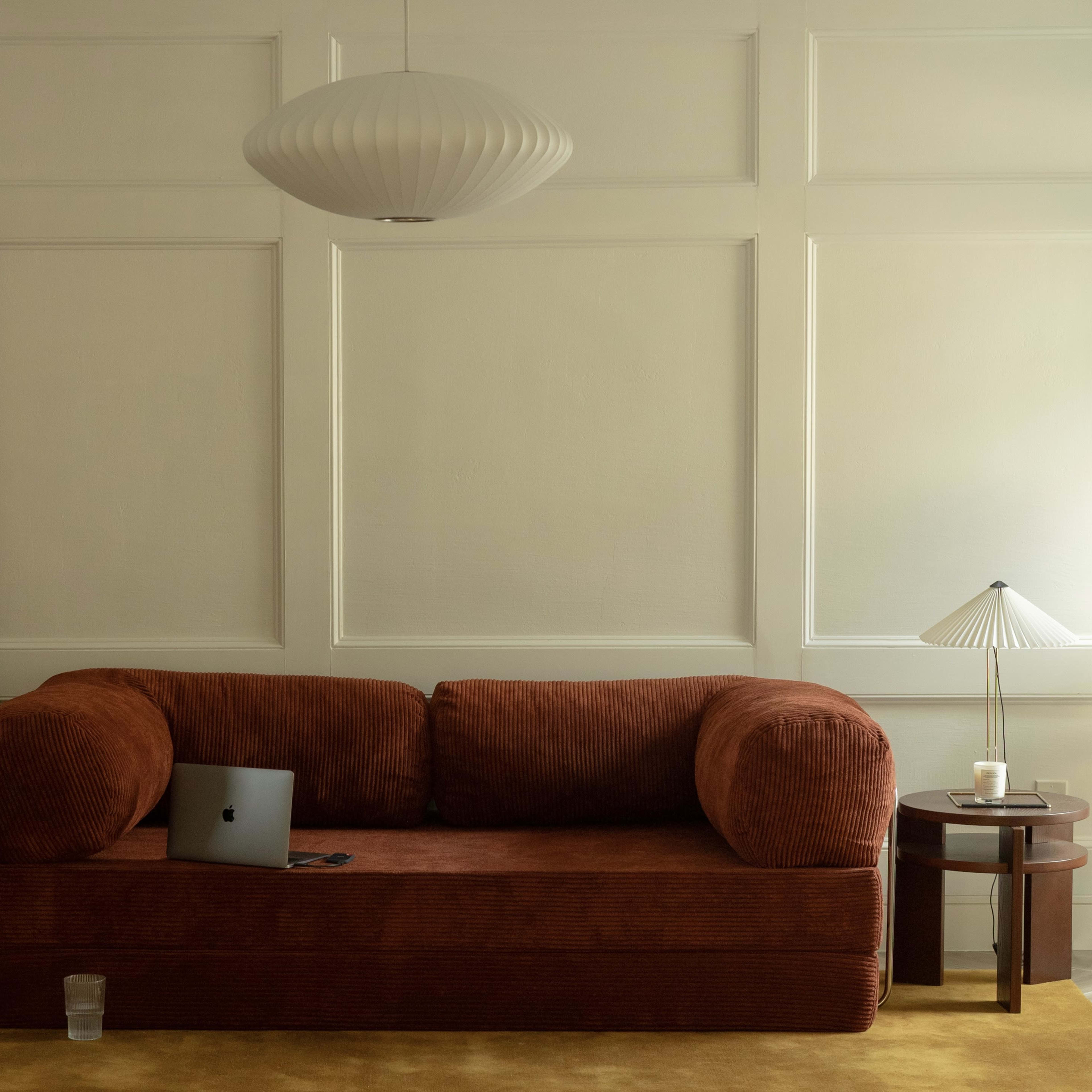 A brown corduroy sofa sits against a wall with paneling, accompanied by a small round table with a lamp and a glass of water nearby.
