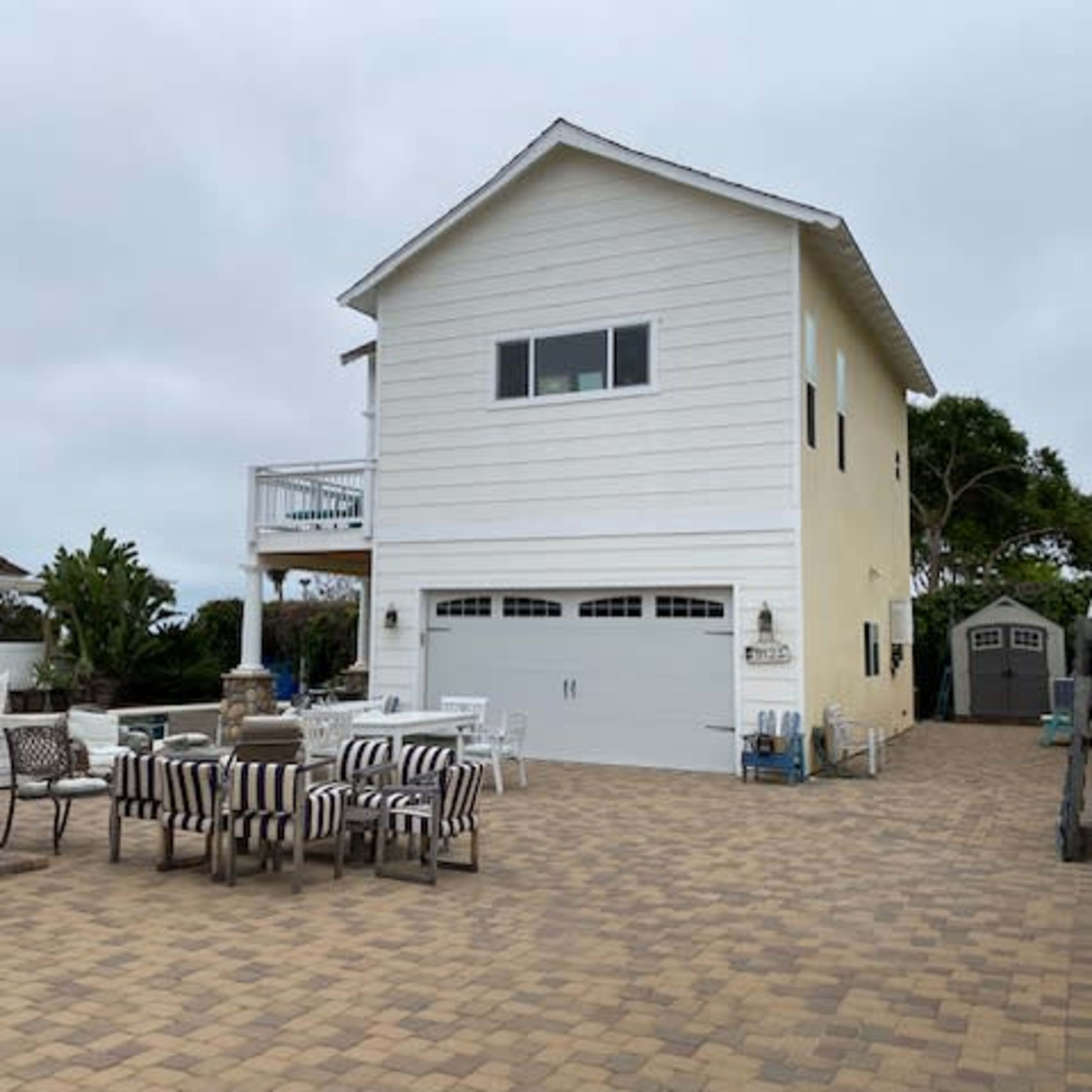A two-story house with a white exterior and a garage is situated on a paved patio area surrounded by outdoor furniture.