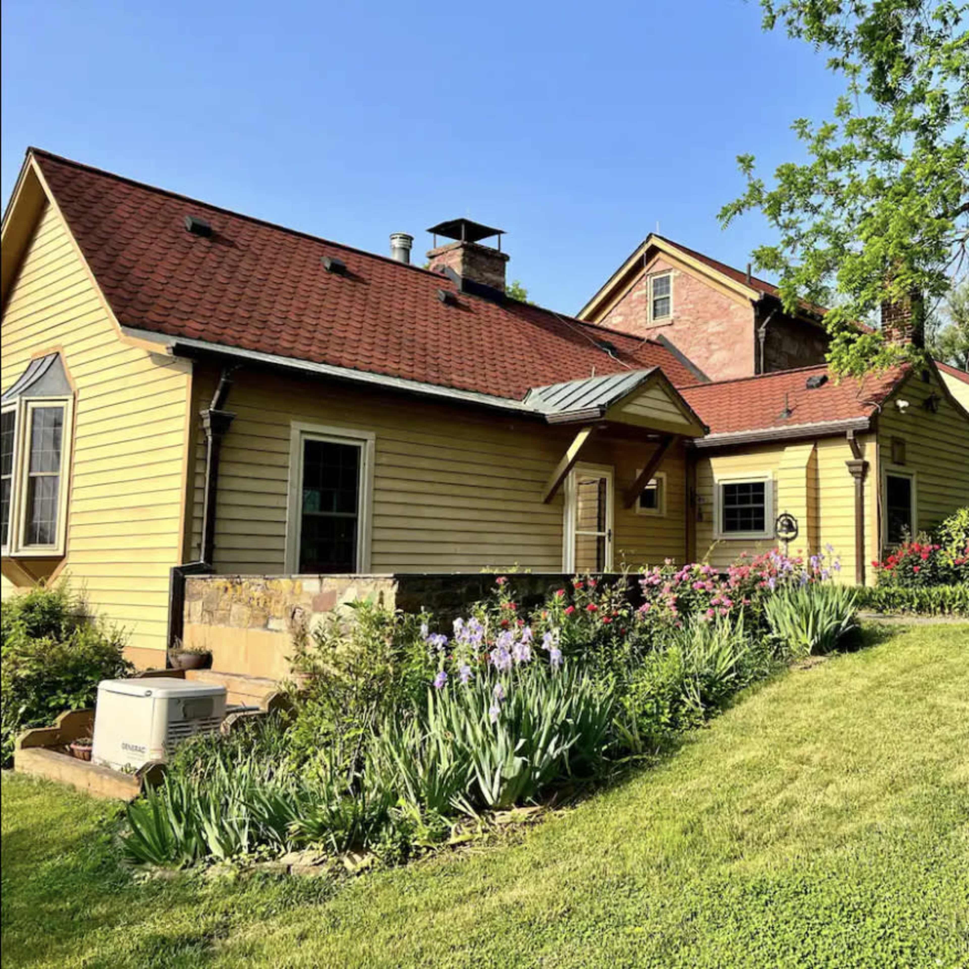 A yellow farmhouse with a red roof is surrounded by green grass and flower beds under a clear blue sky.