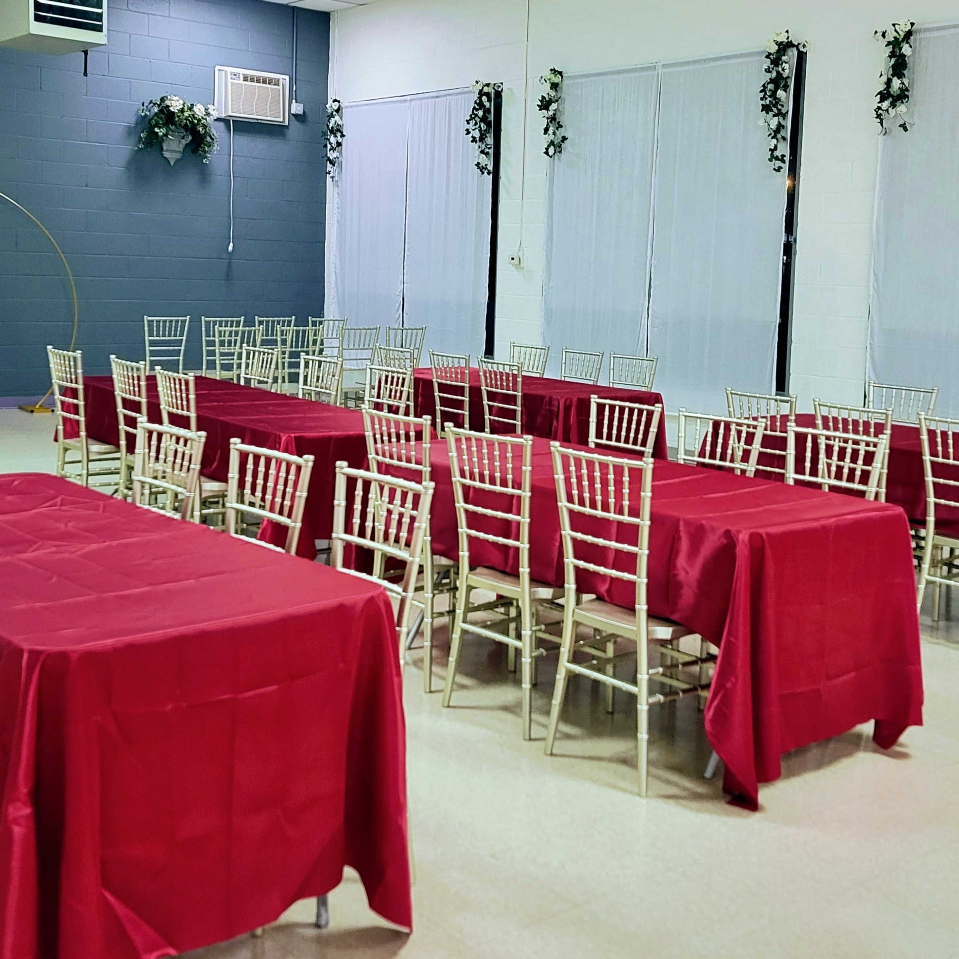 A hall with multiple tables covered in red tablecloths and gold chairs arranged in a grid pattern.