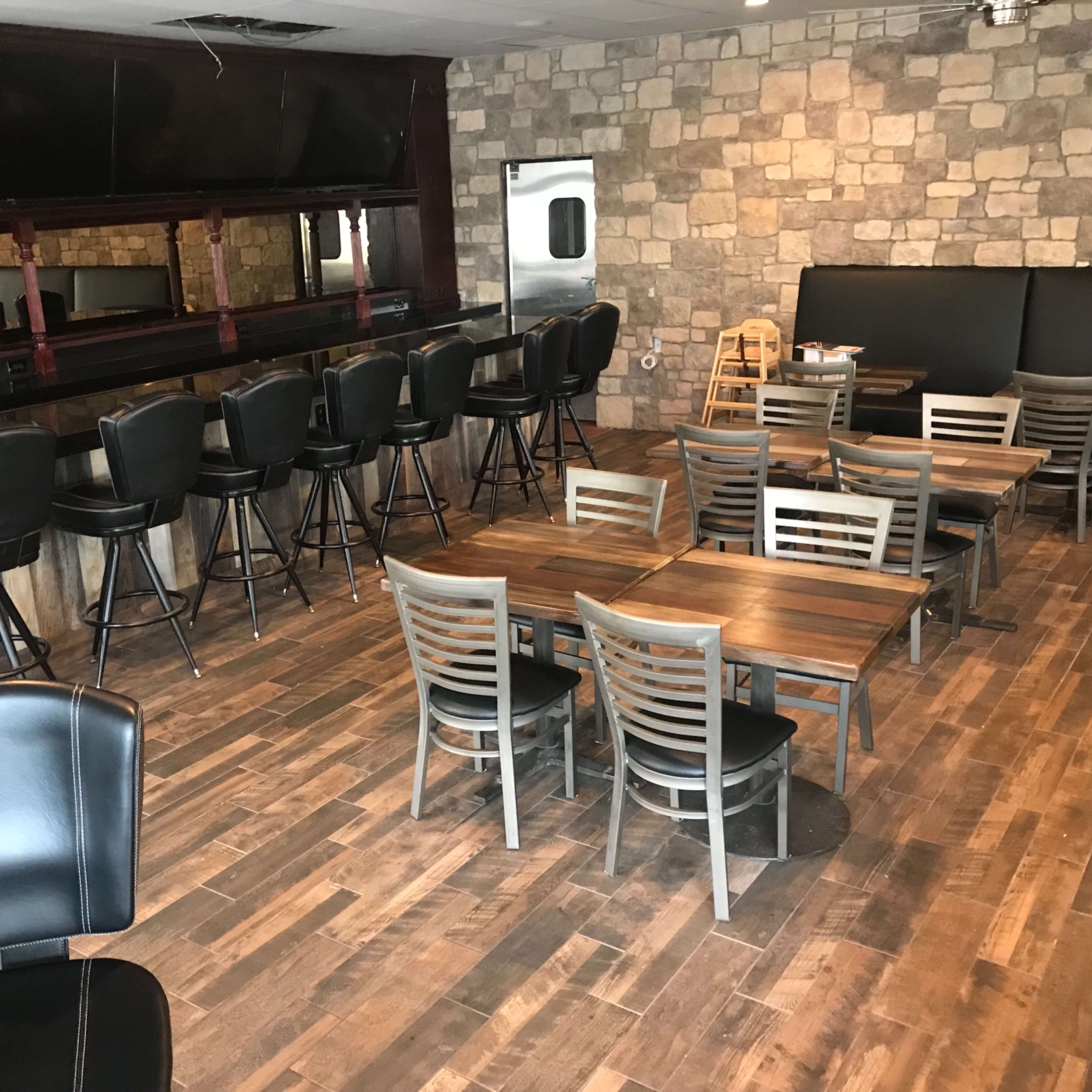 A restaurant interior featuring a bar with high stools, several tables with chairs, and a stone-patterned wall.
