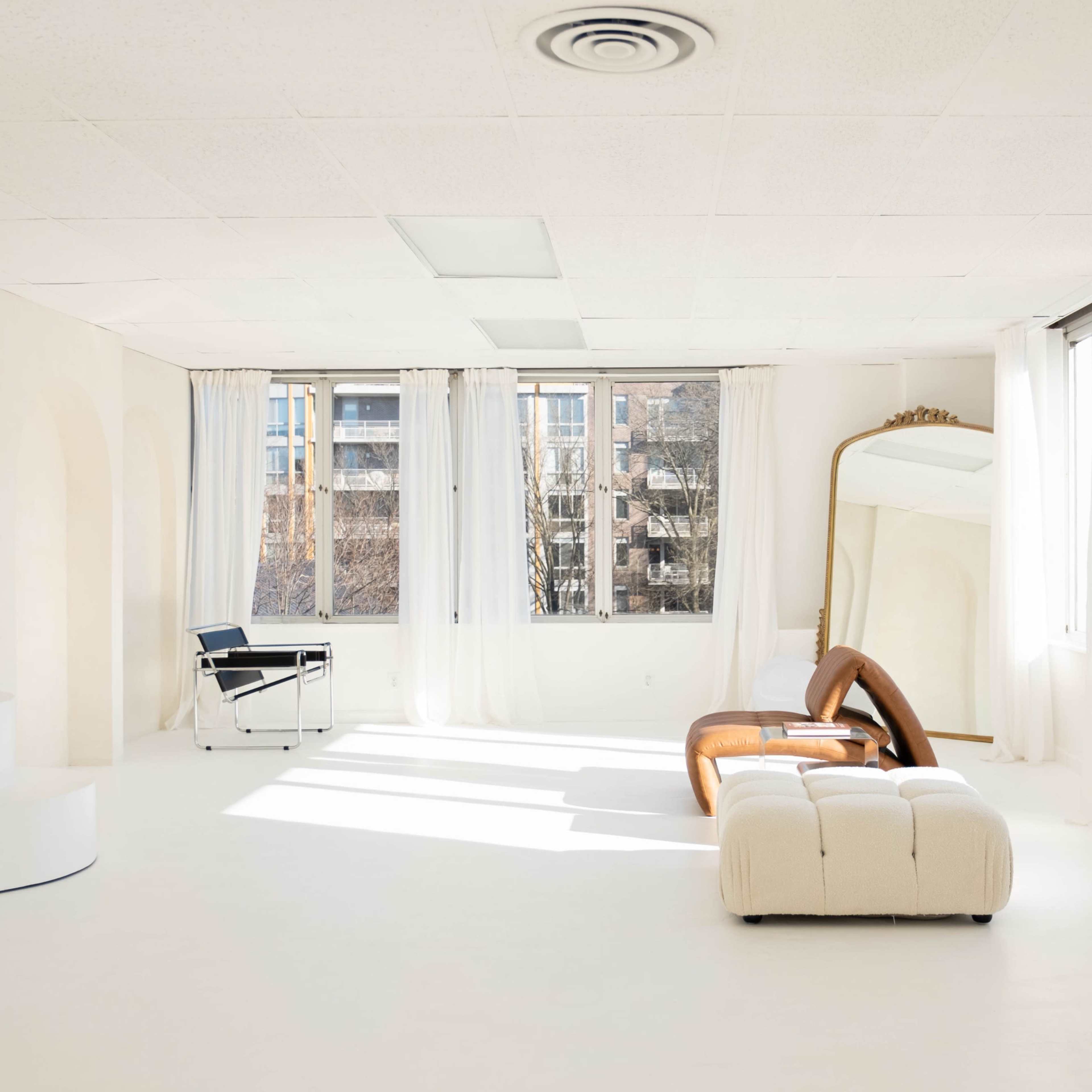 A bright, minimalist room features large windows, white walls, and modern furniture, including a curved sofa and a full-length mirror.