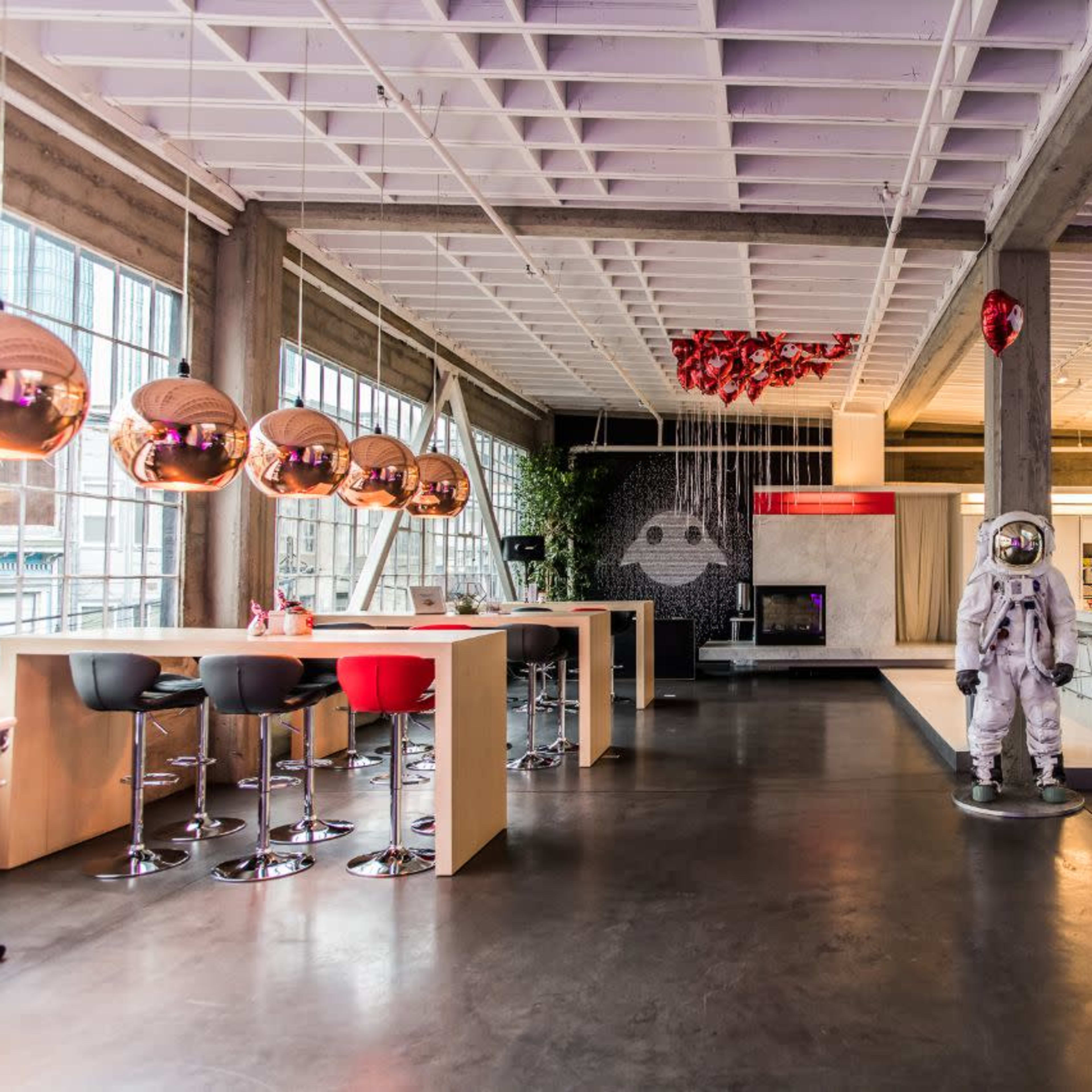 A spacious modern interior features a kitchen area, dining bar with stools, and a life-sized astronaut statue against a backdrop of large windows.
