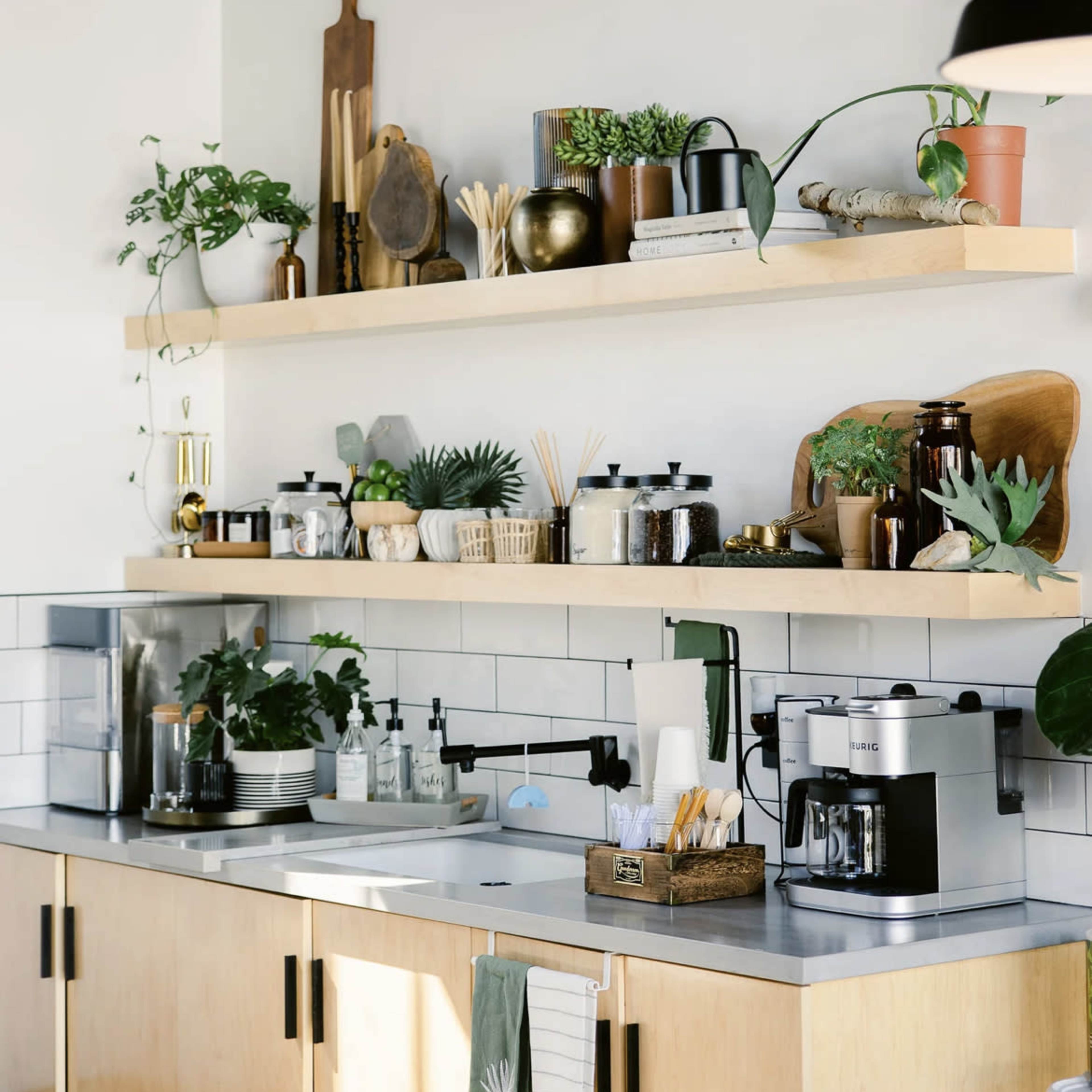 A modern kitchen features wooden shelves above a countertop, adorned with various plants, kitchenware, and glass containers.