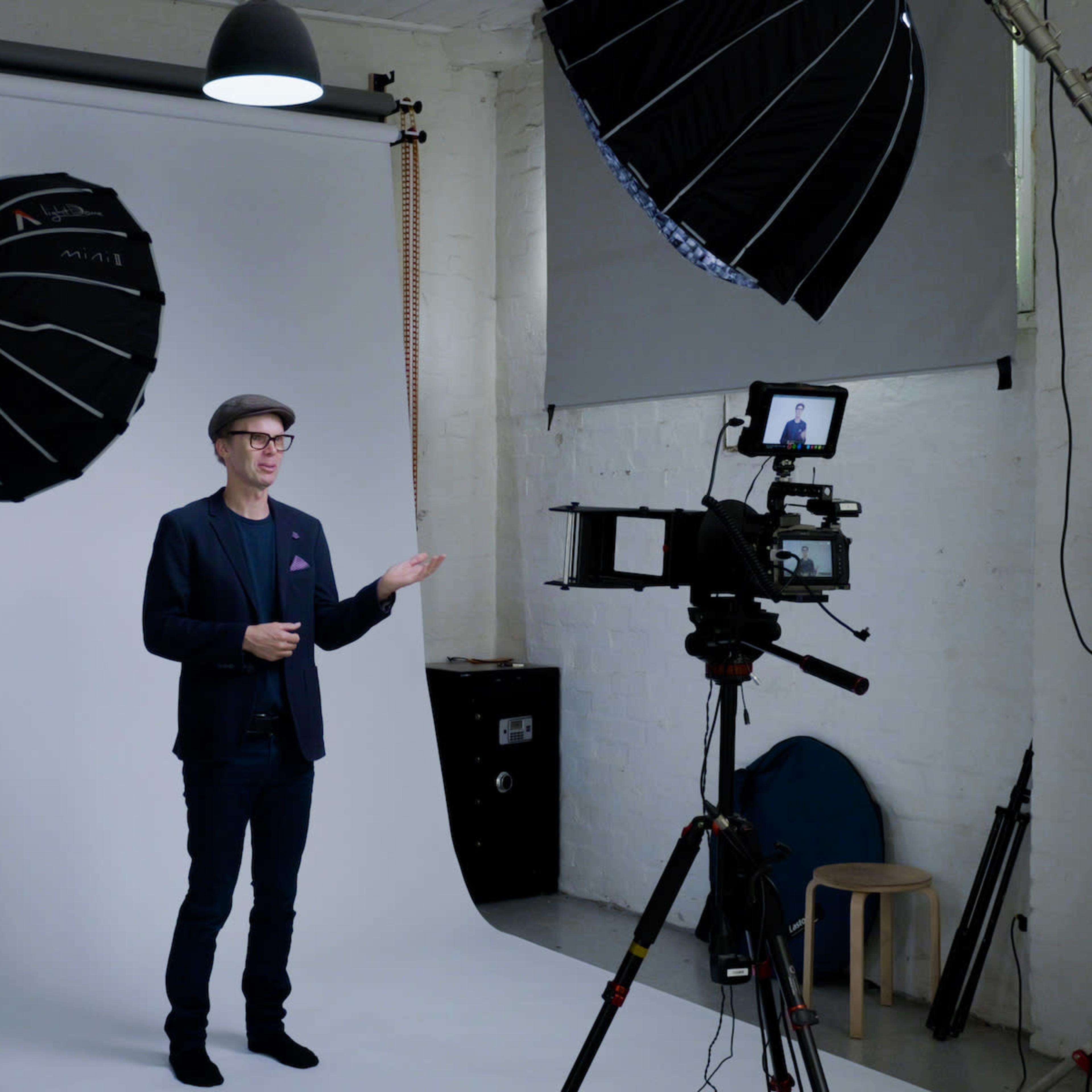 A man stands in front of a backdrop in a photography studio, gesturing towards a camera mounted on a tripod.