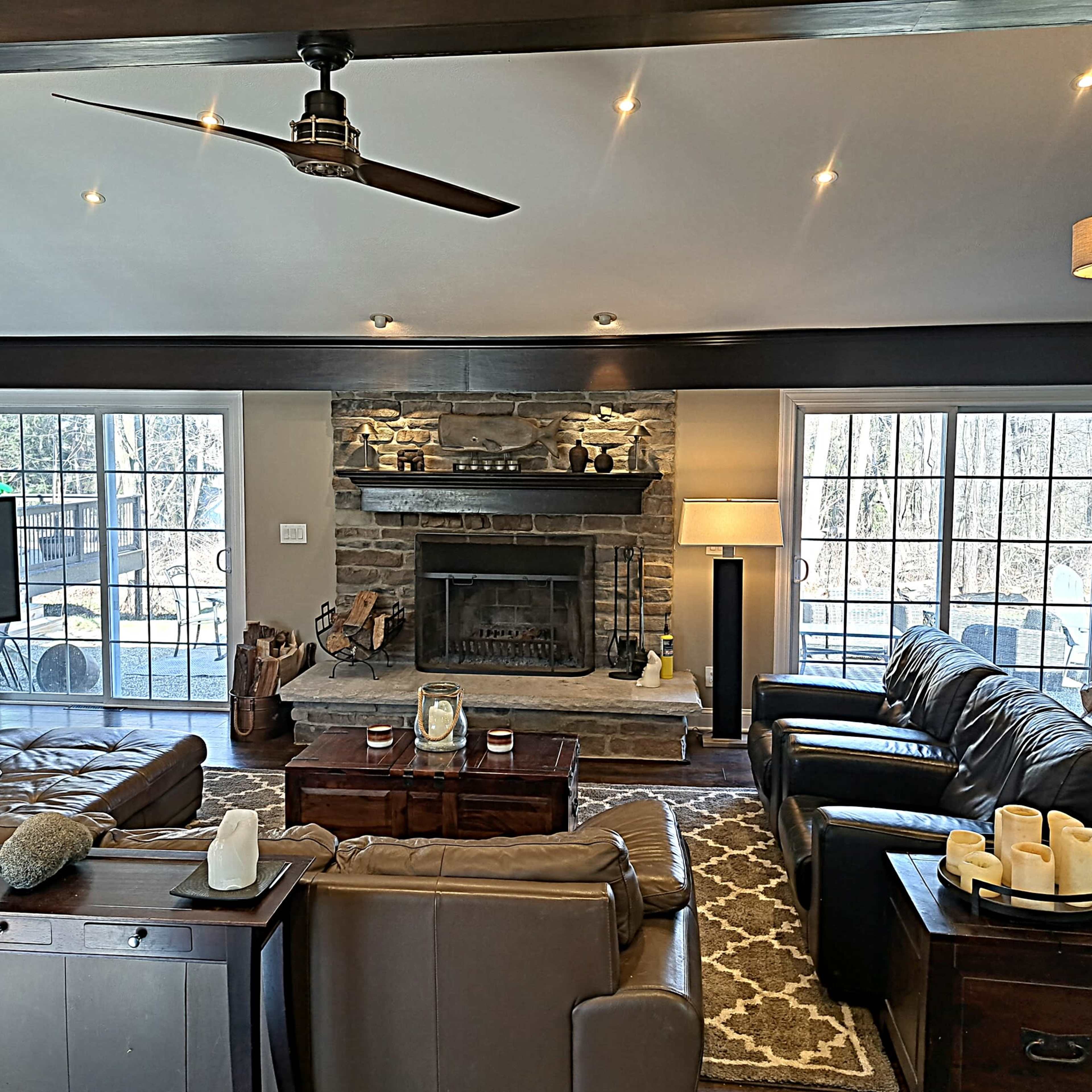 A cozy living room features a stone fireplace, two brown leather sofas, a wooden coffee table, and large windows that provide natural light.
