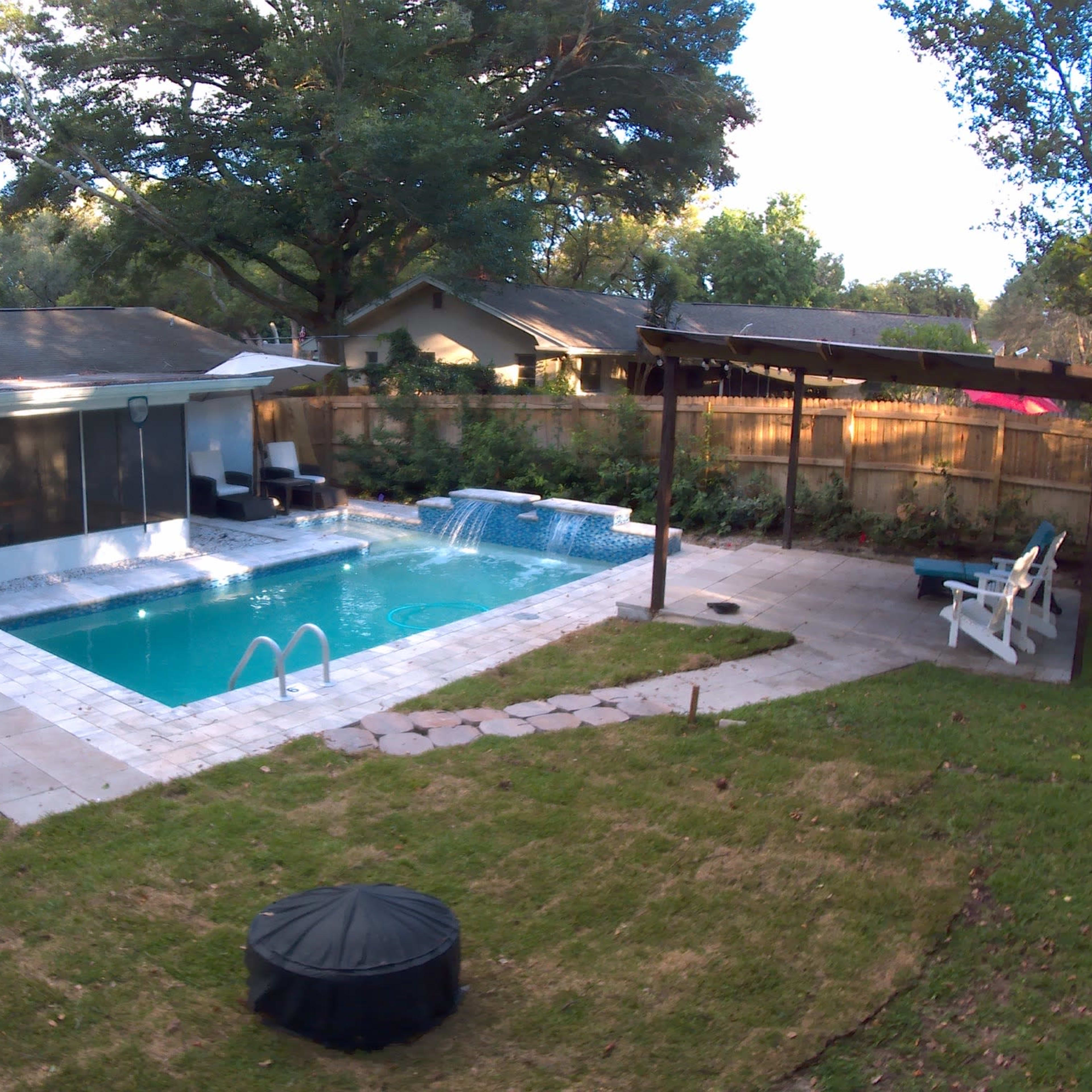 A backyard pool area with a swimming pool, two lounge chairs, and a pergola surrounded by green grass and plants.