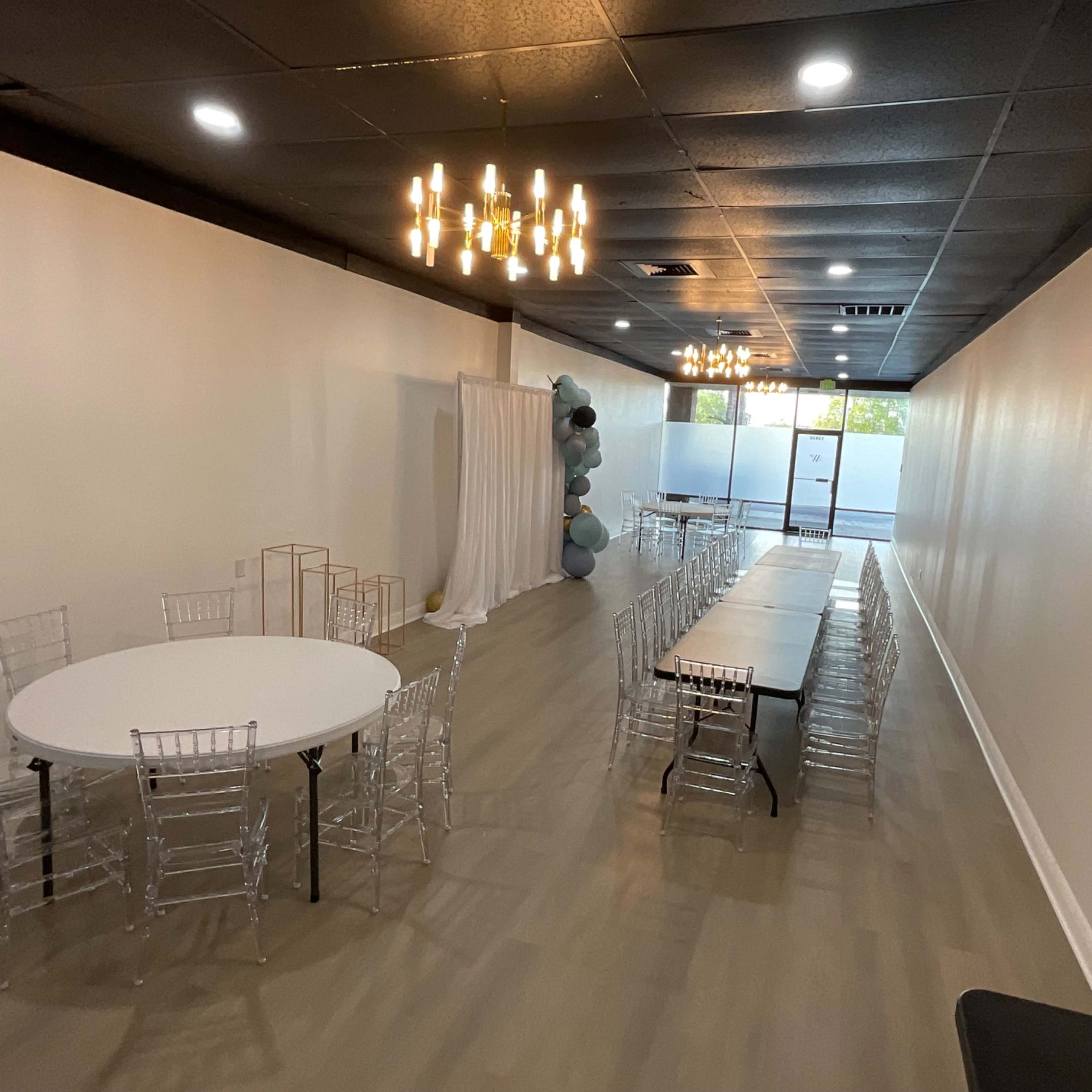 A spacious event room with a round table and a long rectangular table set up, along with decorative elements and another area for a backdrop.