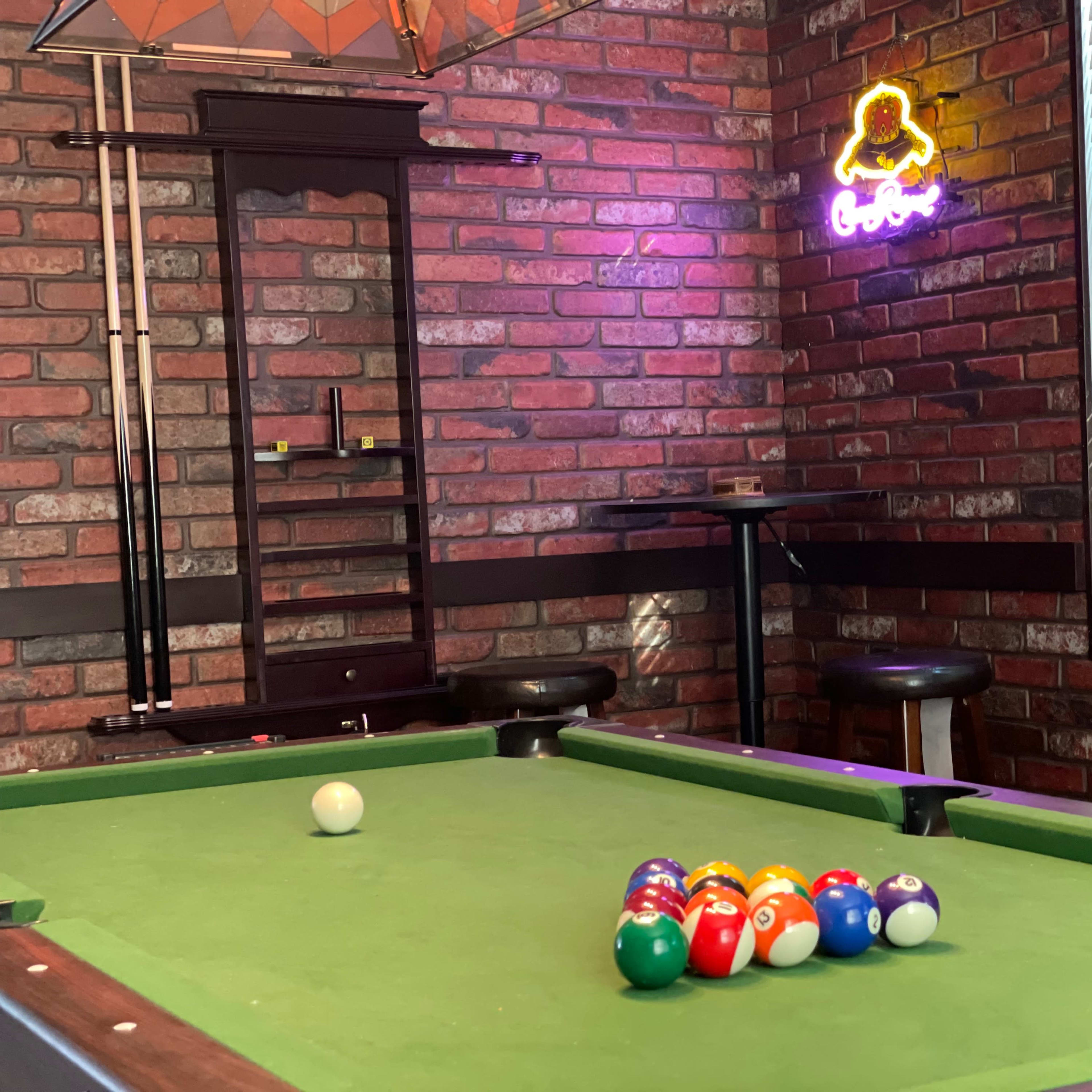 A pool table with colorful balls arranged in a triangle sits in a room with brick walls and a neon sign.