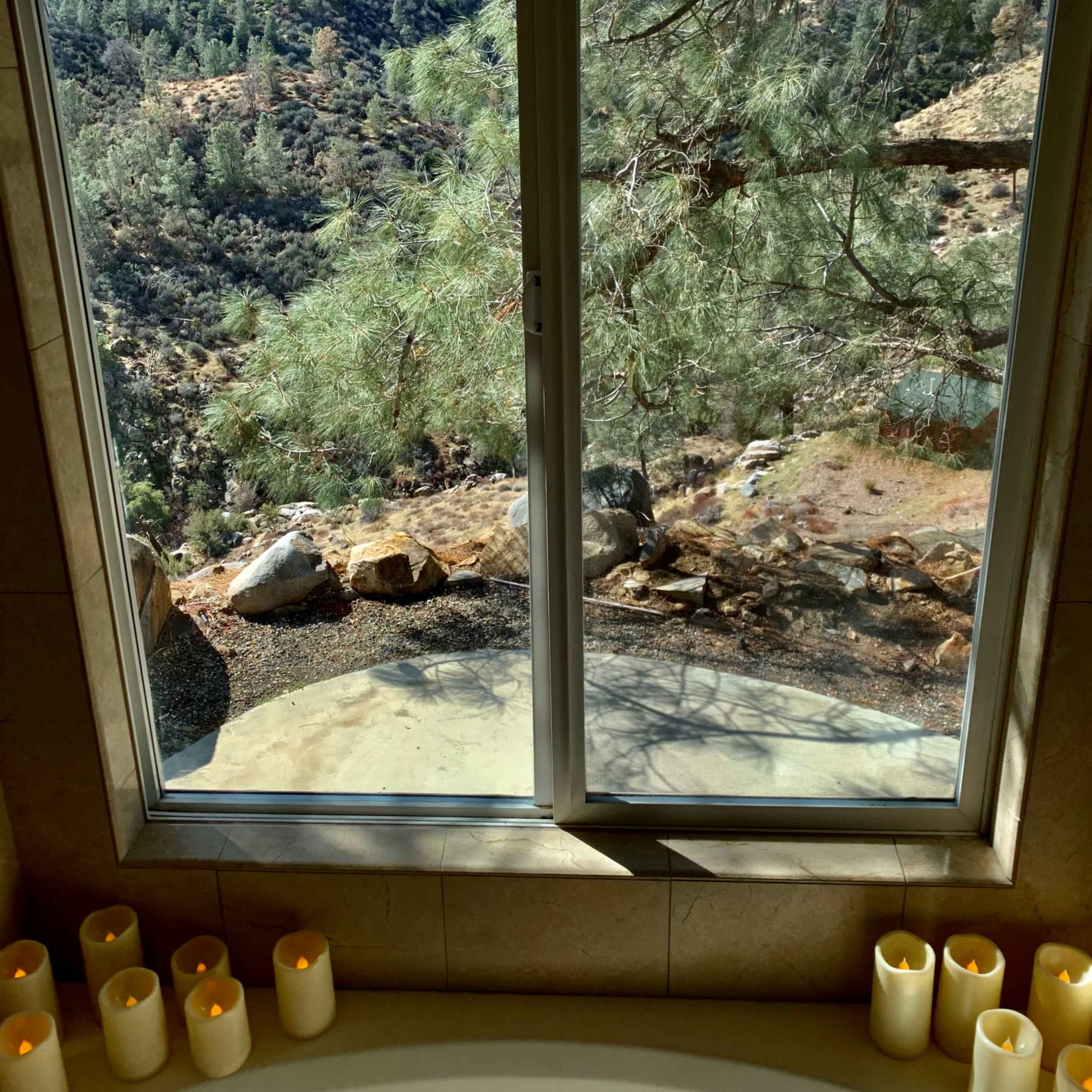 A view from a bathroom window reveals a rocky landscape and trees outside, with candles lined up along the edge of a bathtub.