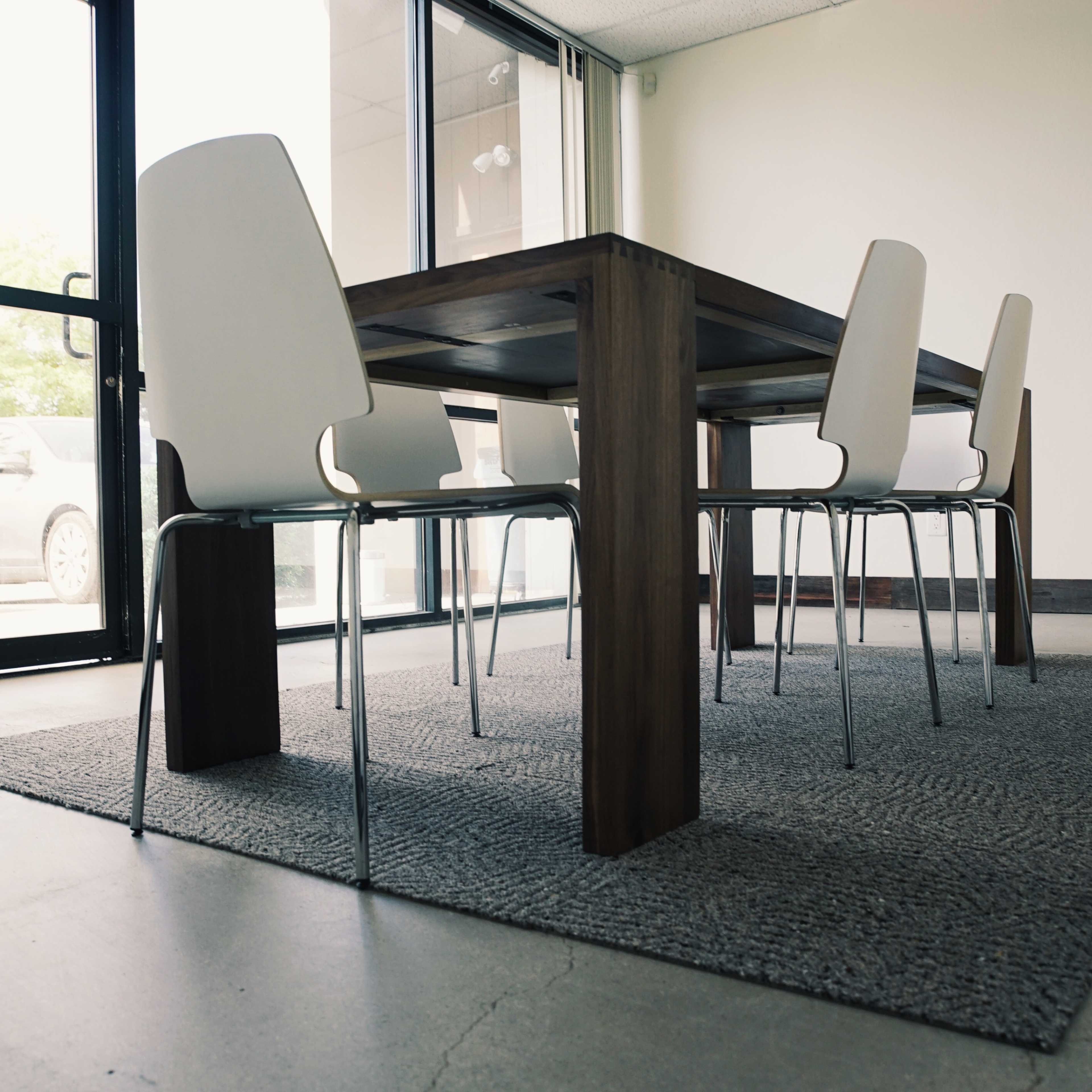A wooden table with four metal chairs sits on a gray rug in a spacious, well-lit room with large windows.