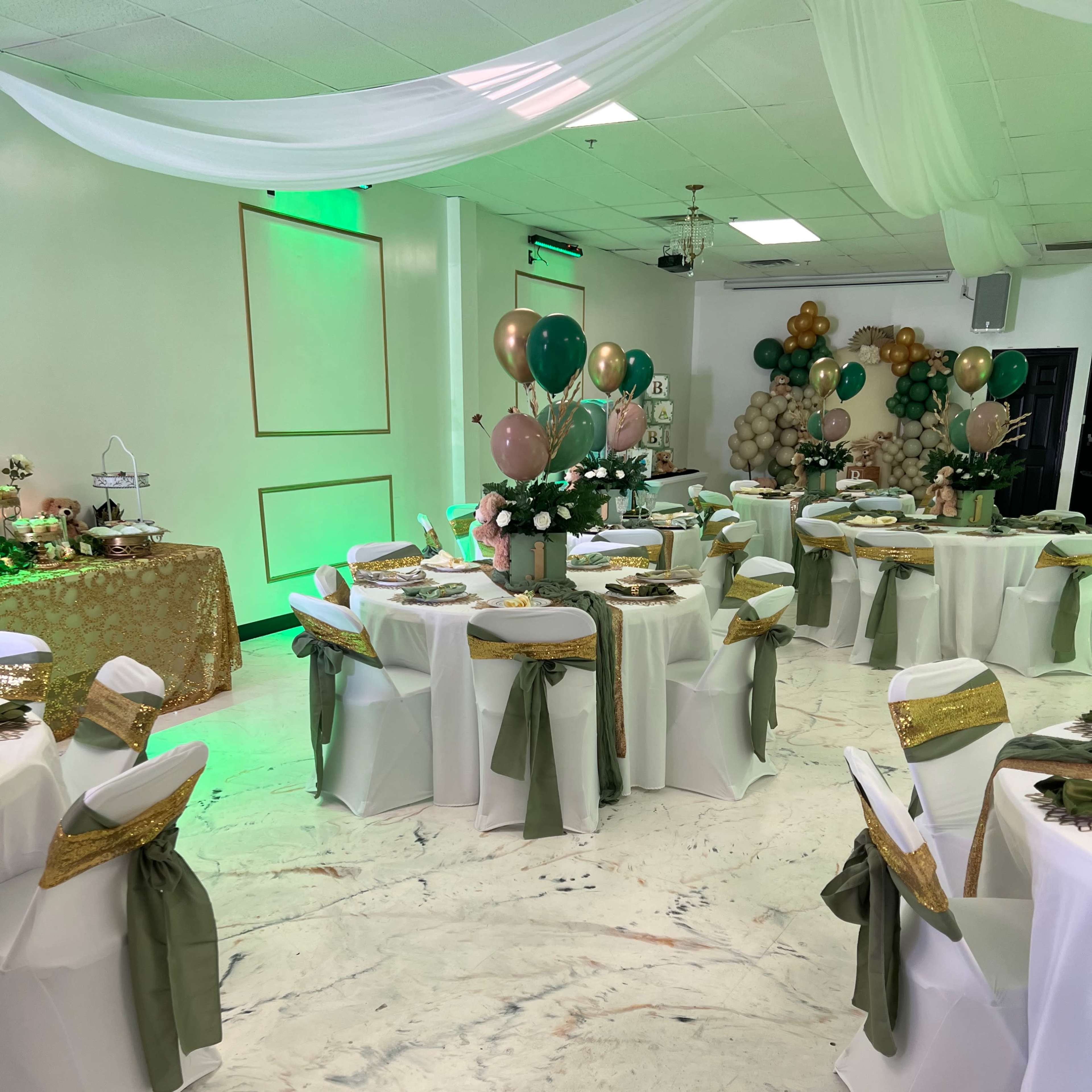 A banquet room set up for an event, featuring round tables with white tablecloths, green sashes, and decorative centerpieces, surrounded by balloons in gold, green, and pink.