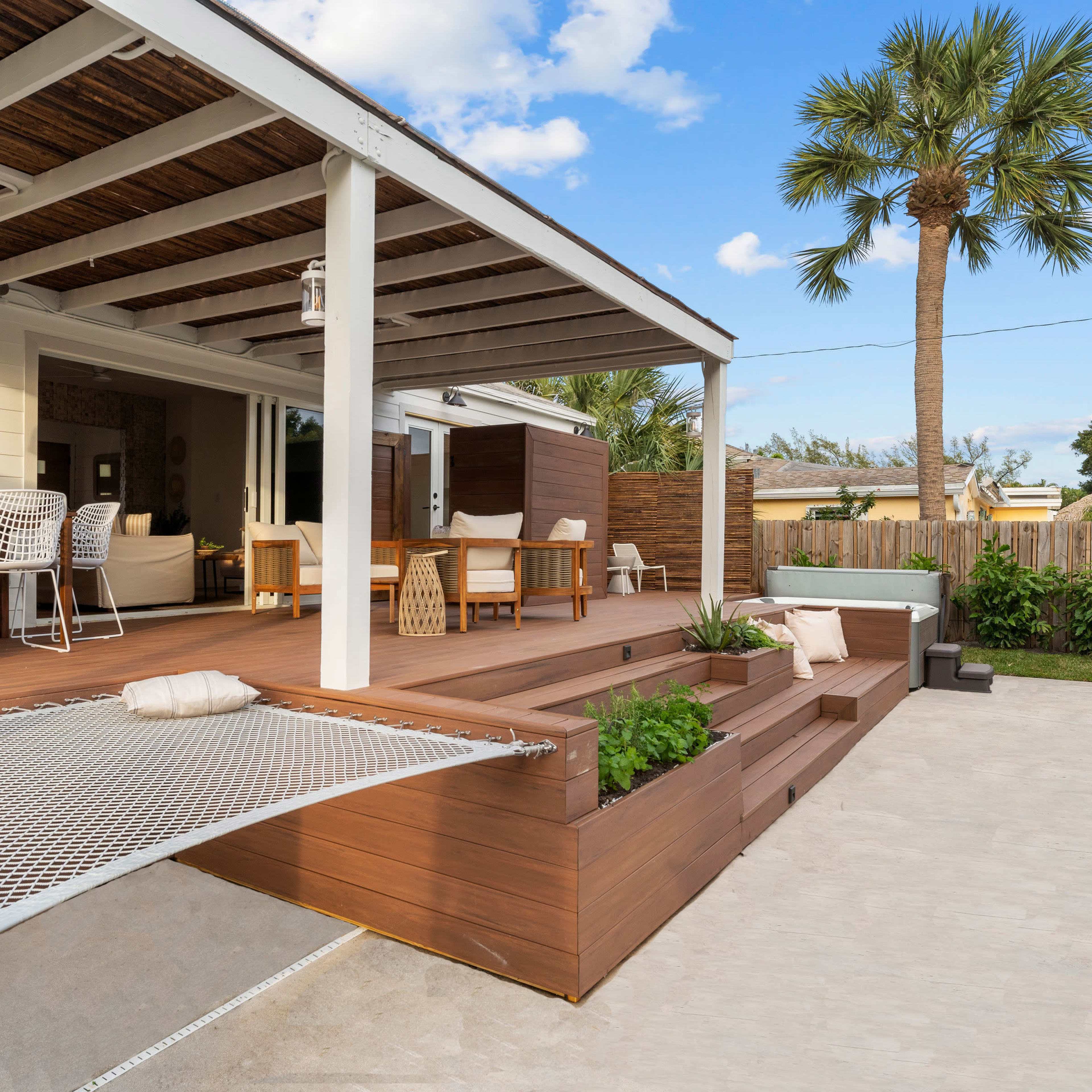 A wooden deck with lounge seating, a dining area, and palm trees surrounds a backyard patio with a hammock and planters.