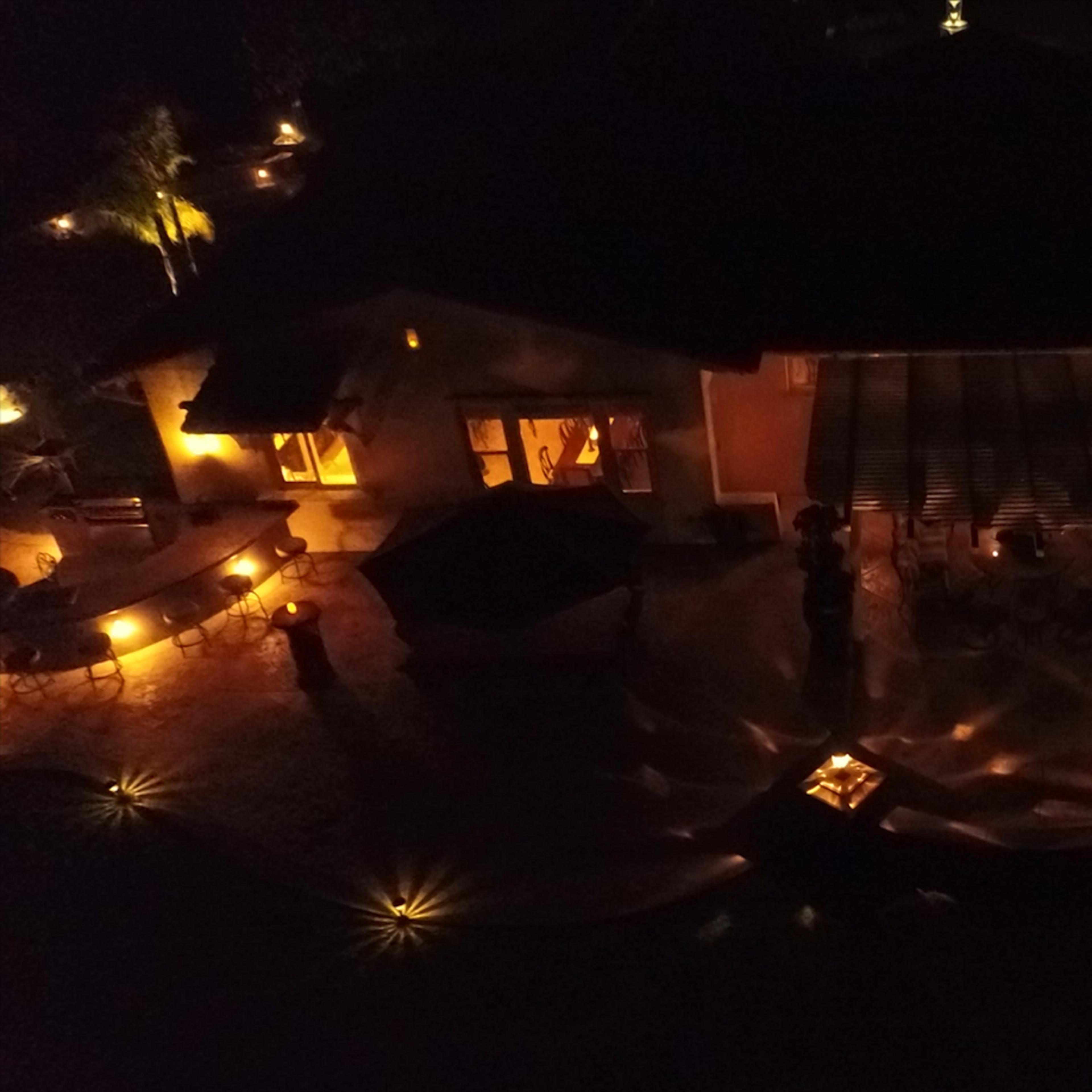 A well-lit outdoor gathering is taking place around a house, surrounded by illuminated pathways and soft lighting in the evening.