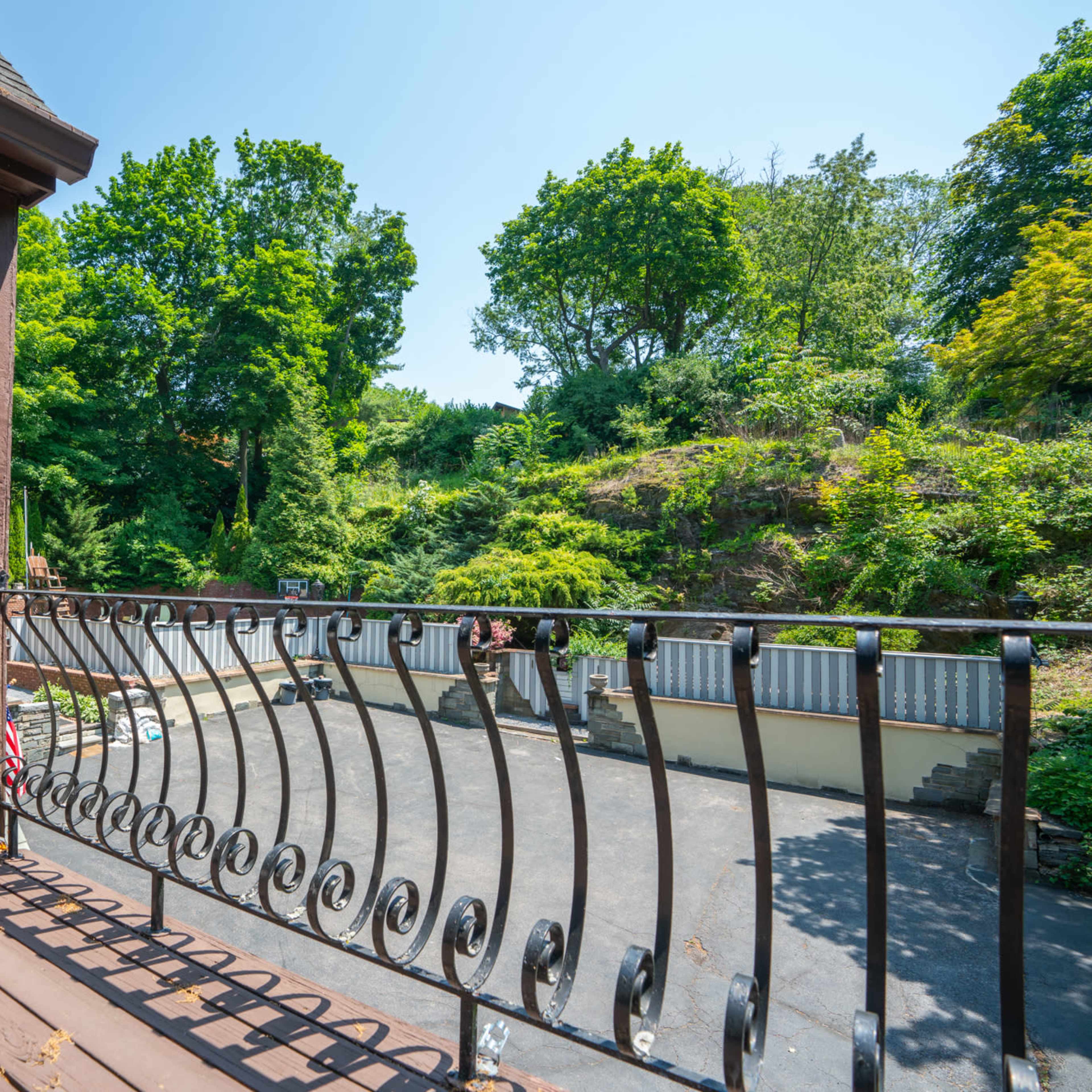 A balcony railing overlooks a paved area surrounded by trees and greenery.