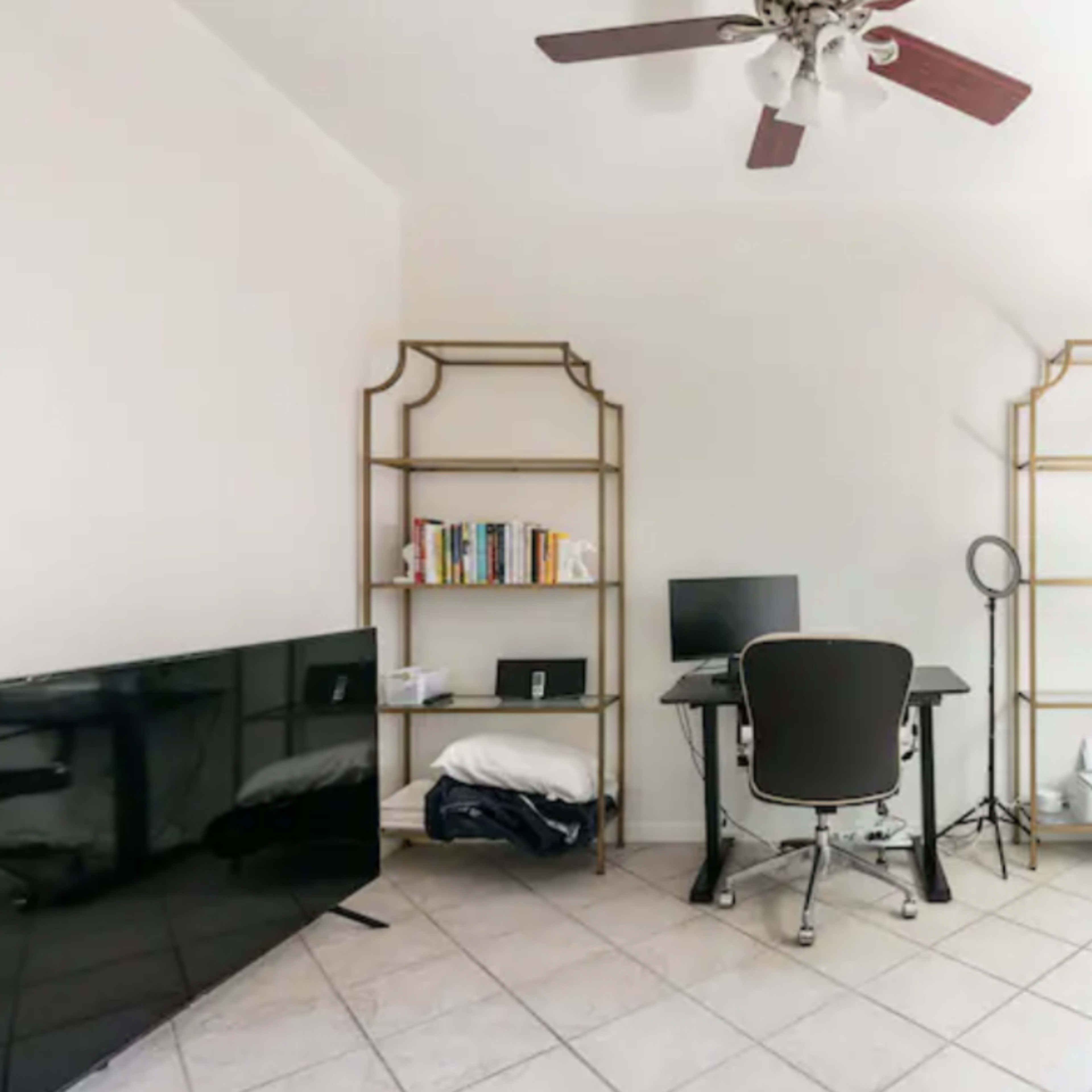 A minimalist home office with a black desk and chair, a computer monitor, a bookshelf filled with books, and a large television against one wall.