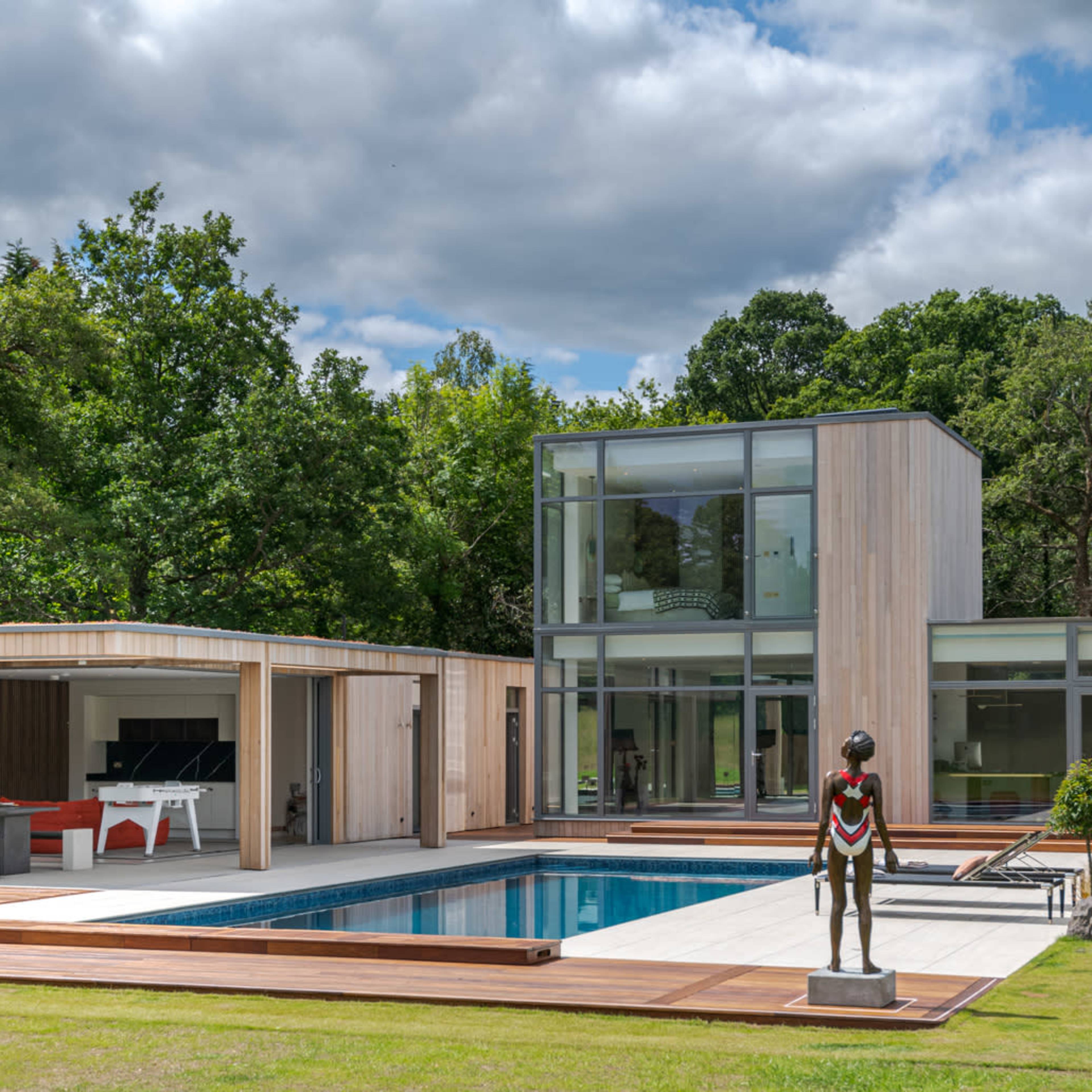 A modern house with large glass windows, a swimming pool, and a sculpture in the foreground, all set in a landscaped area with trees.