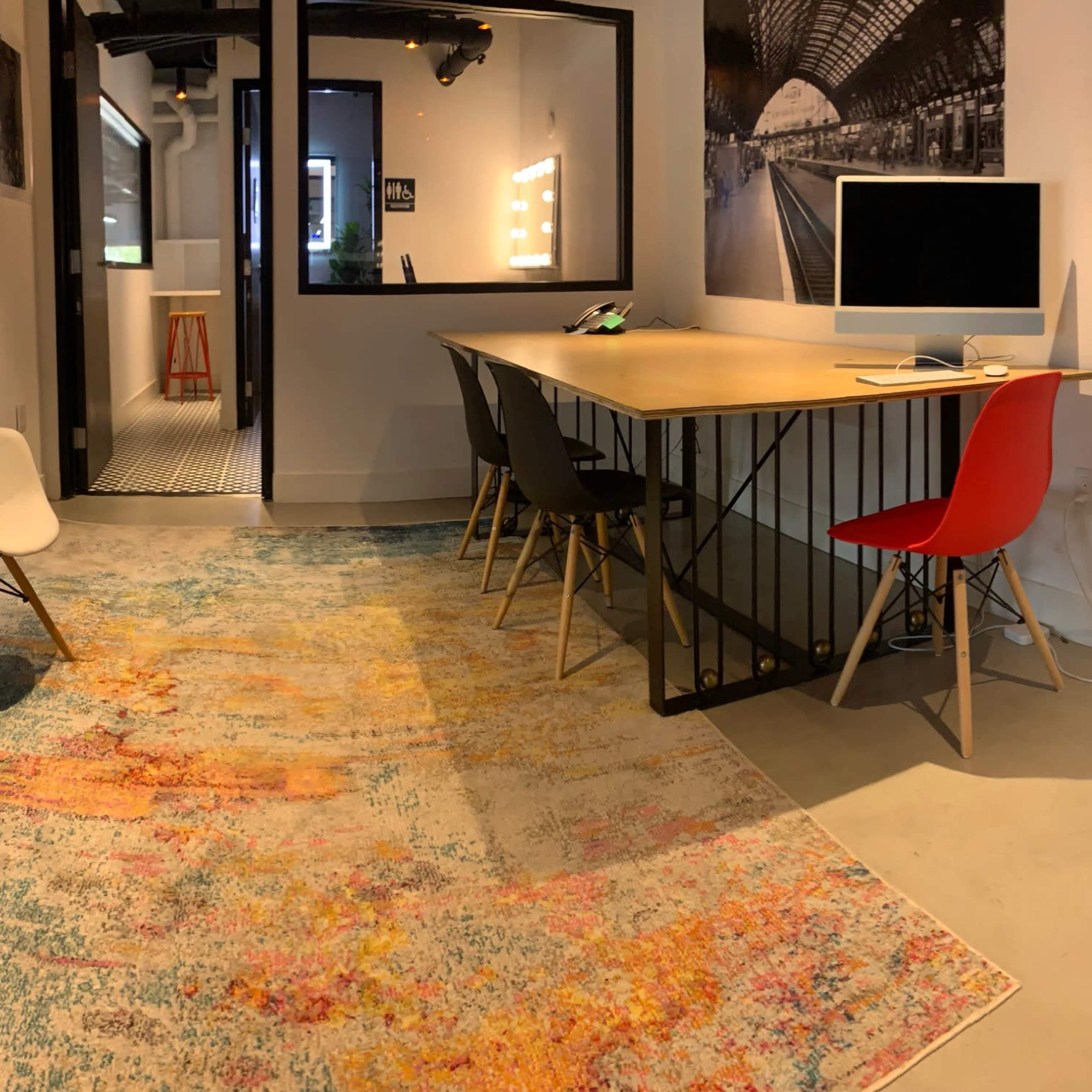 A modern office space with a table surrounded by chairs, a computer on the desk, and a colorful area rug on a concrete floor.