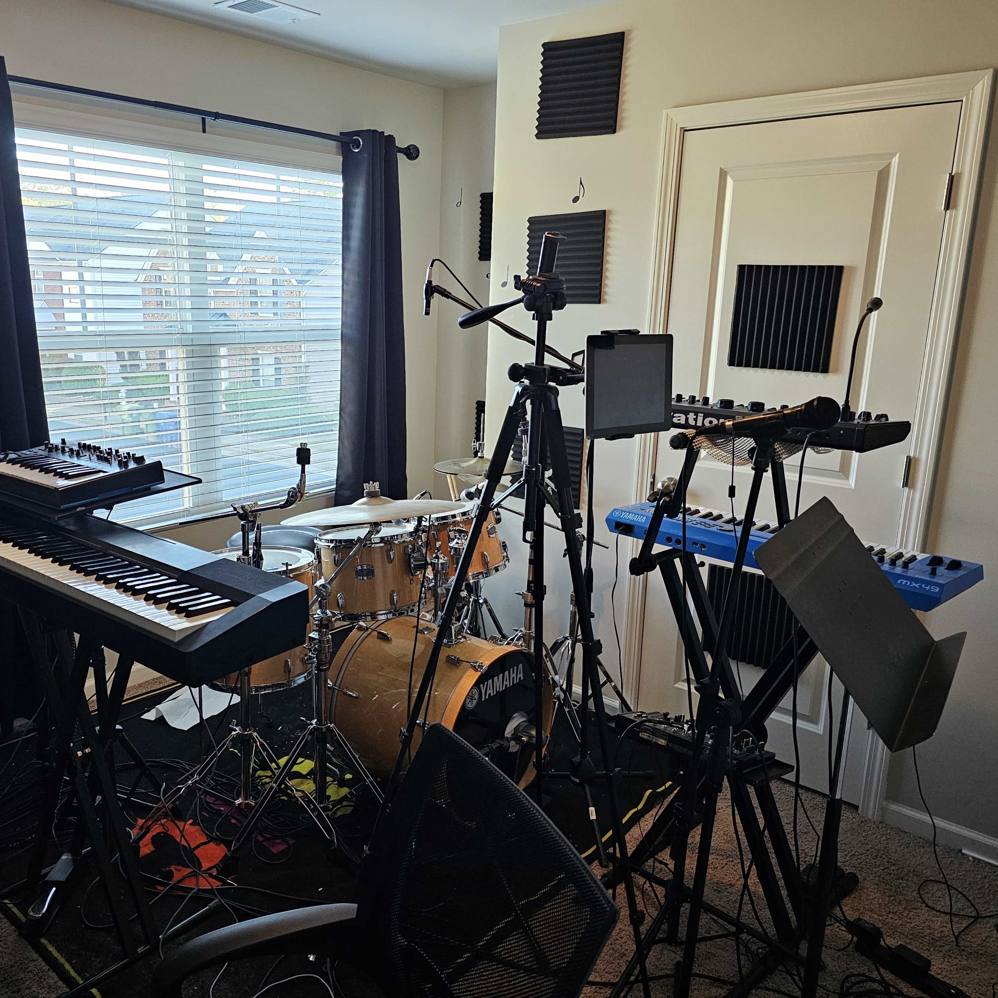 A music studio filled with various musical instruments, including keyboards, a drum set, and audio equipment, in a room with a window and soundproofing panels on the walls.