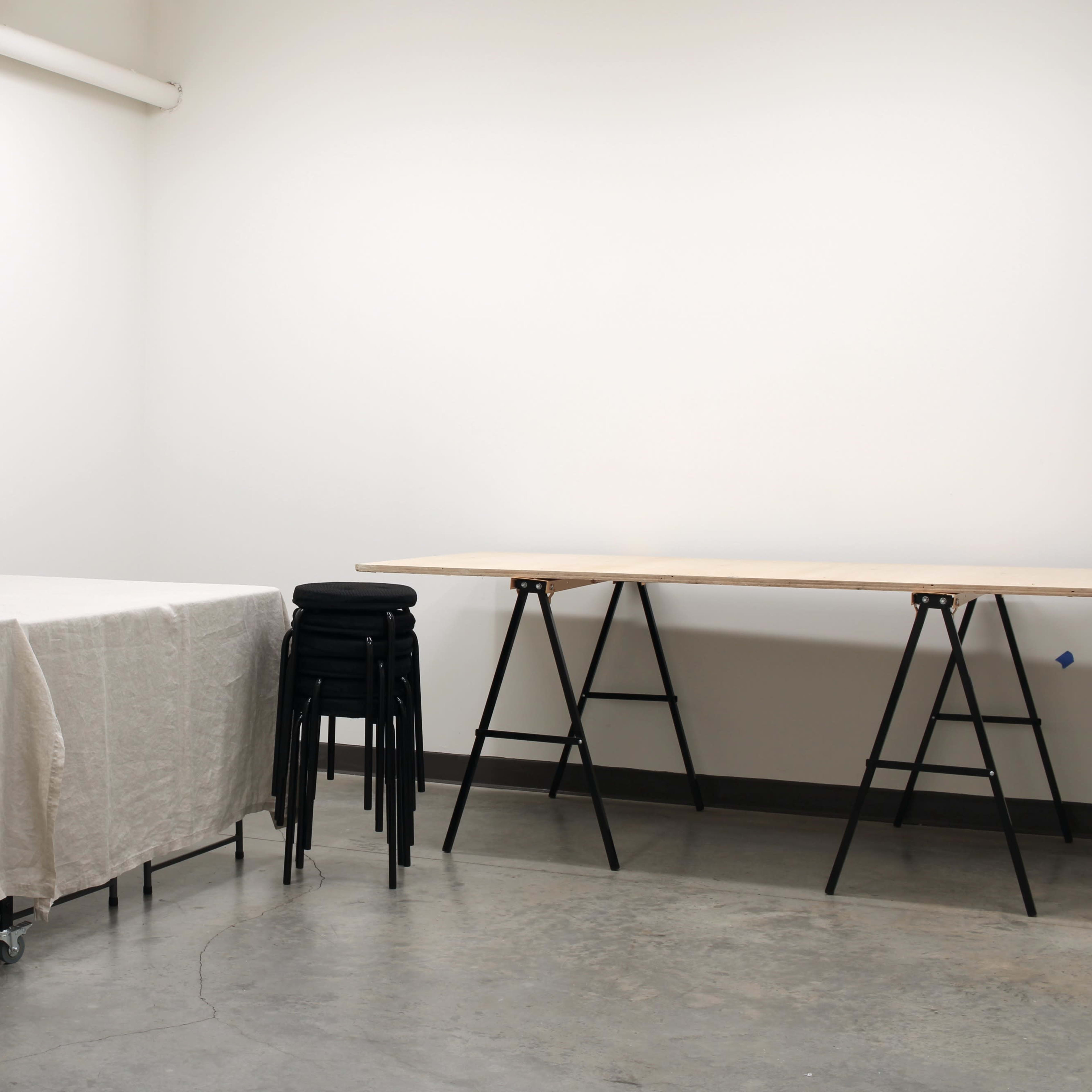 A sparsely furnished room with a table covered in a beige cloth and a stack of black chairs near a wooden table on sawhorse legs.
