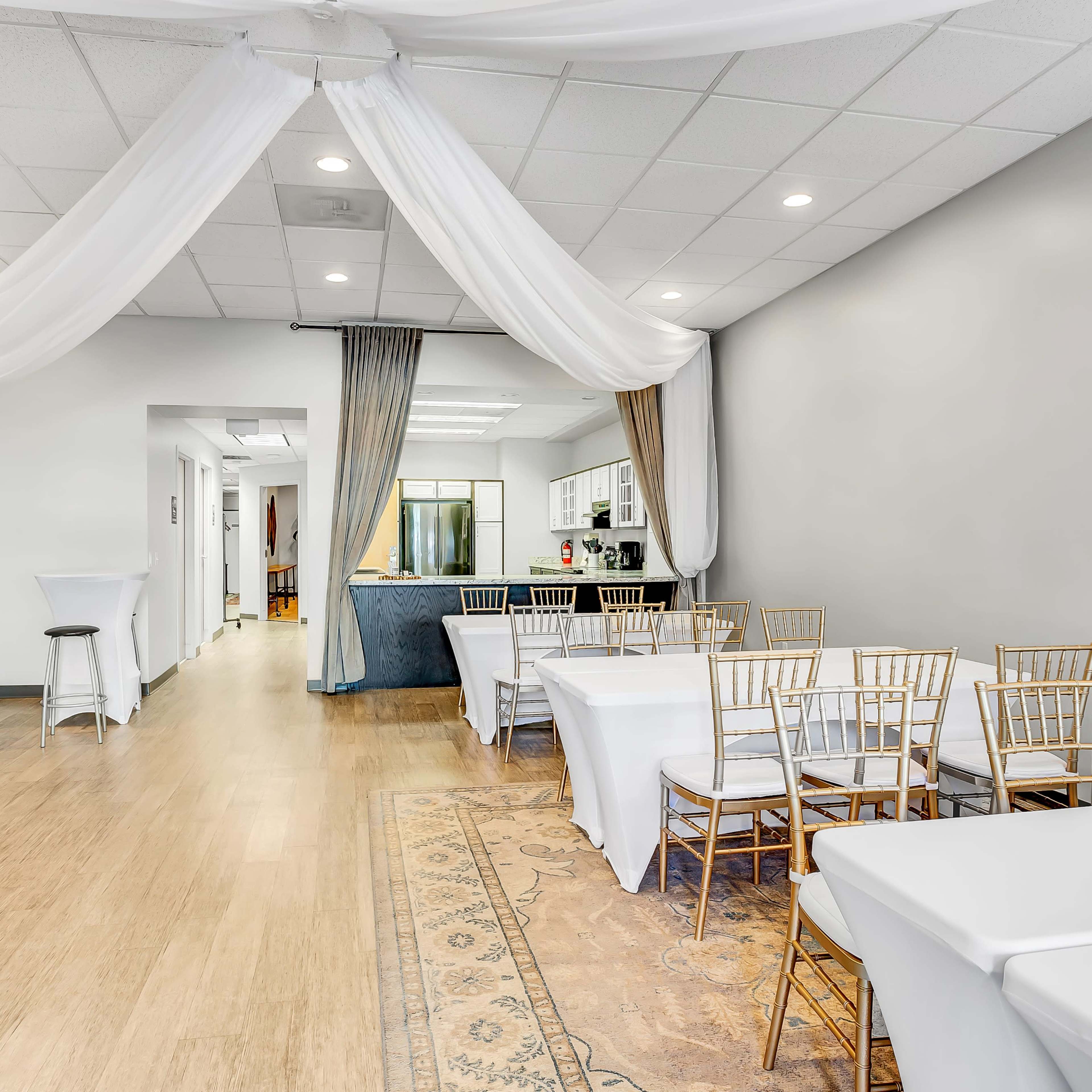 A spacious event room with white tables and chairs arranged for seating, accented by draped fabric overhead and a kitchen area visible in the background.