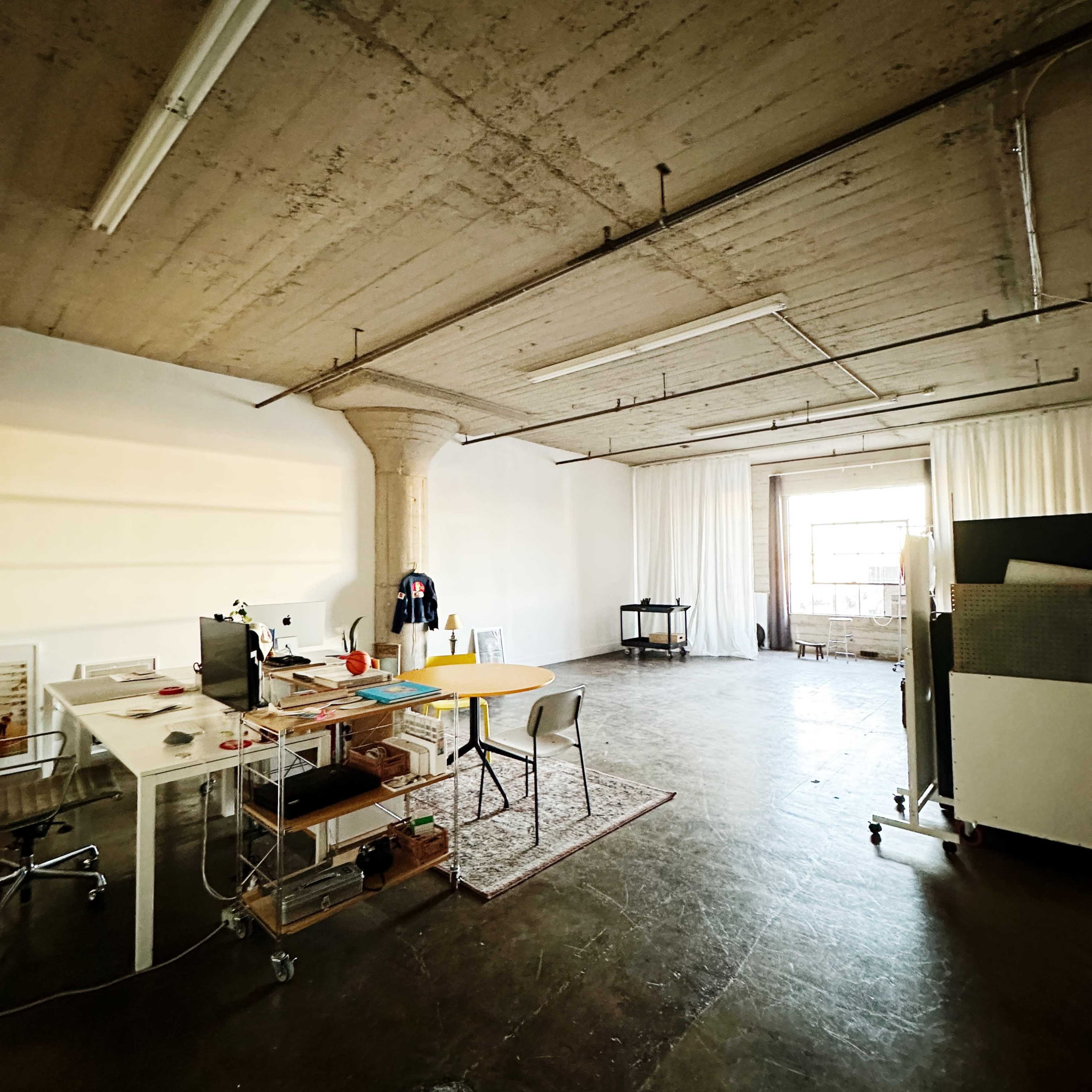 A spacious, industrial-style office with a large window, a desk with a computer, and minimal furnishings.