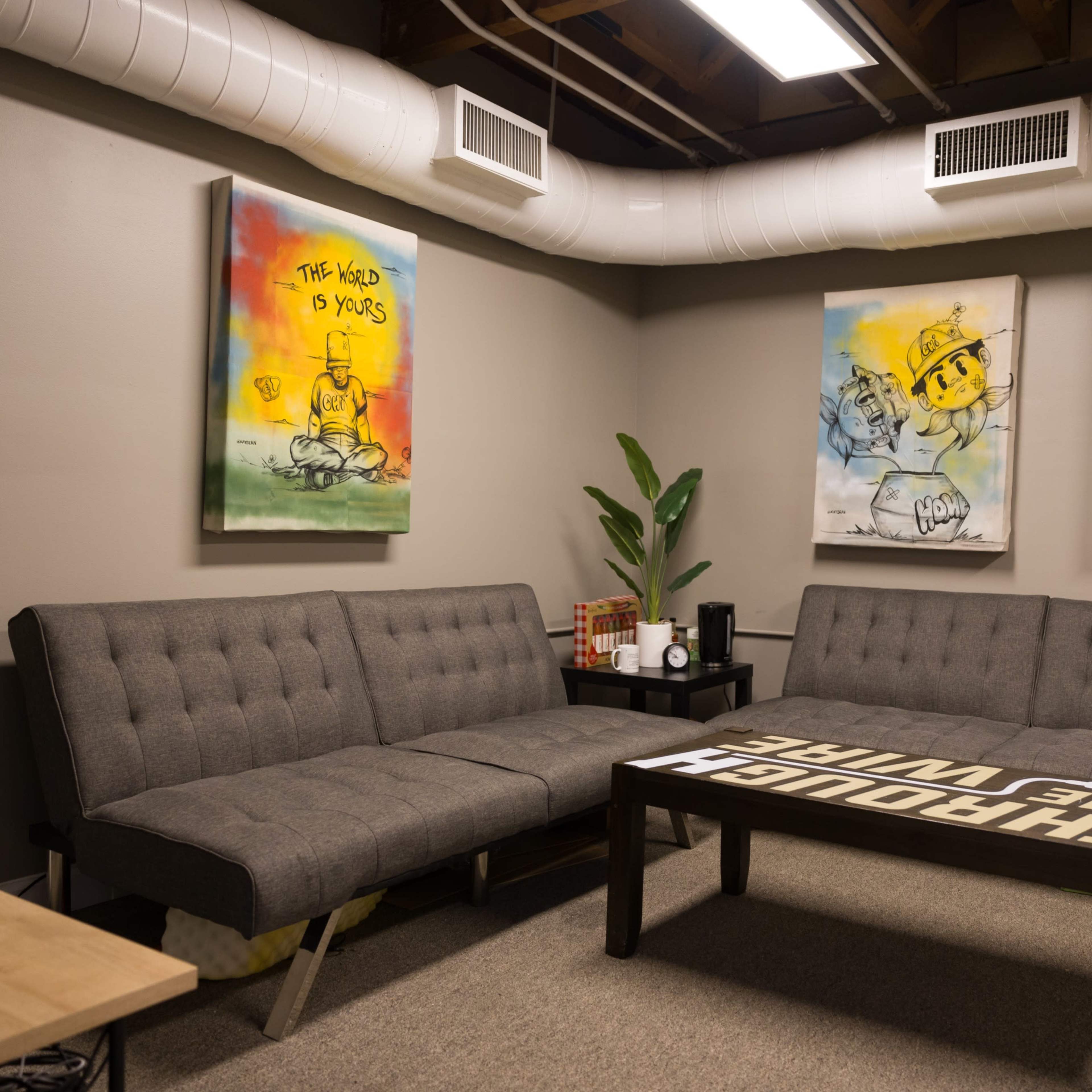 A casual lounge area with a gray sectional sofa, a coffee table printed with the phrase "THROUGH THE WIRE," and three colorful art pieces on the walls.