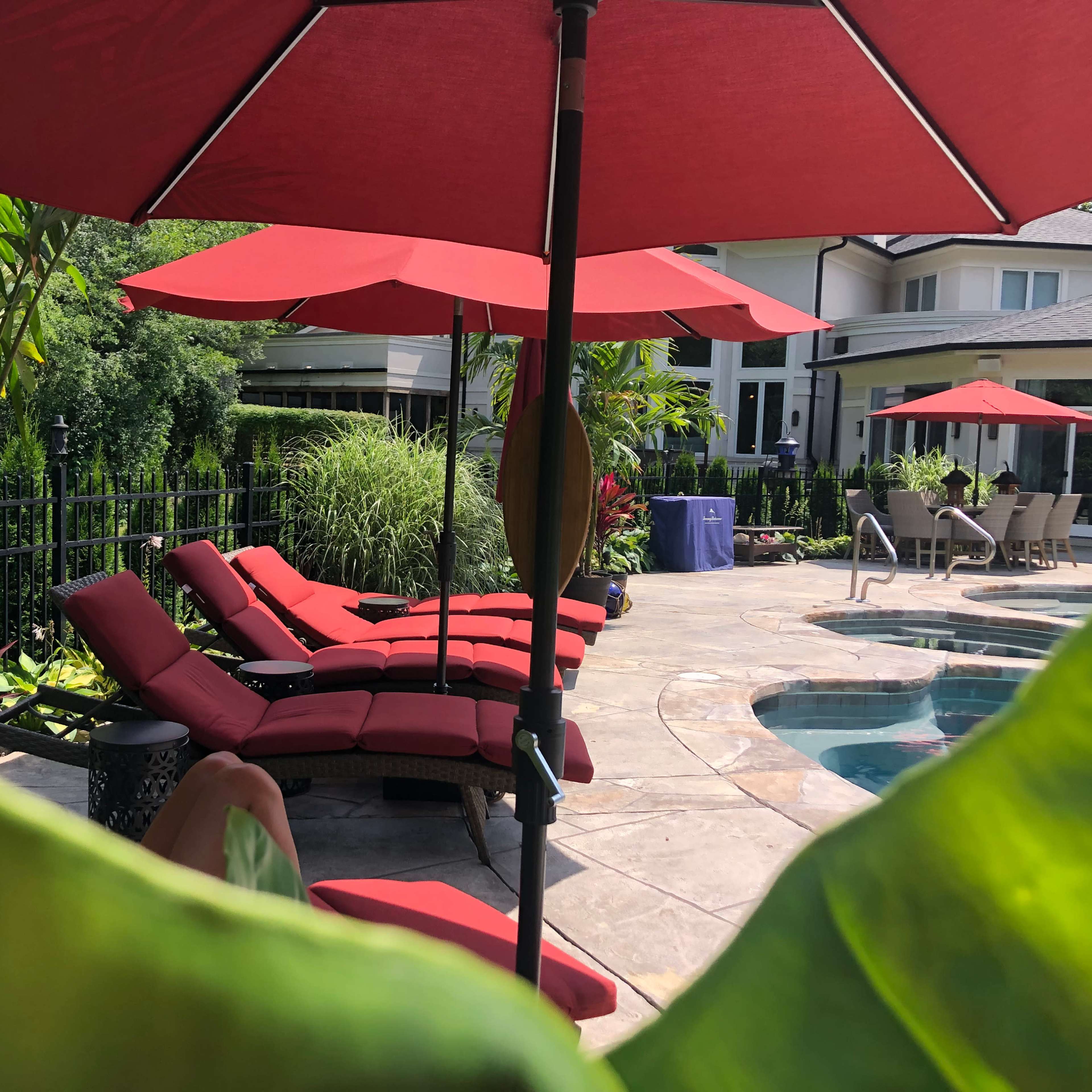 A set of red lounge chairs under matching umbrellas is arranged around a swimming pool in a landscaped backyard.