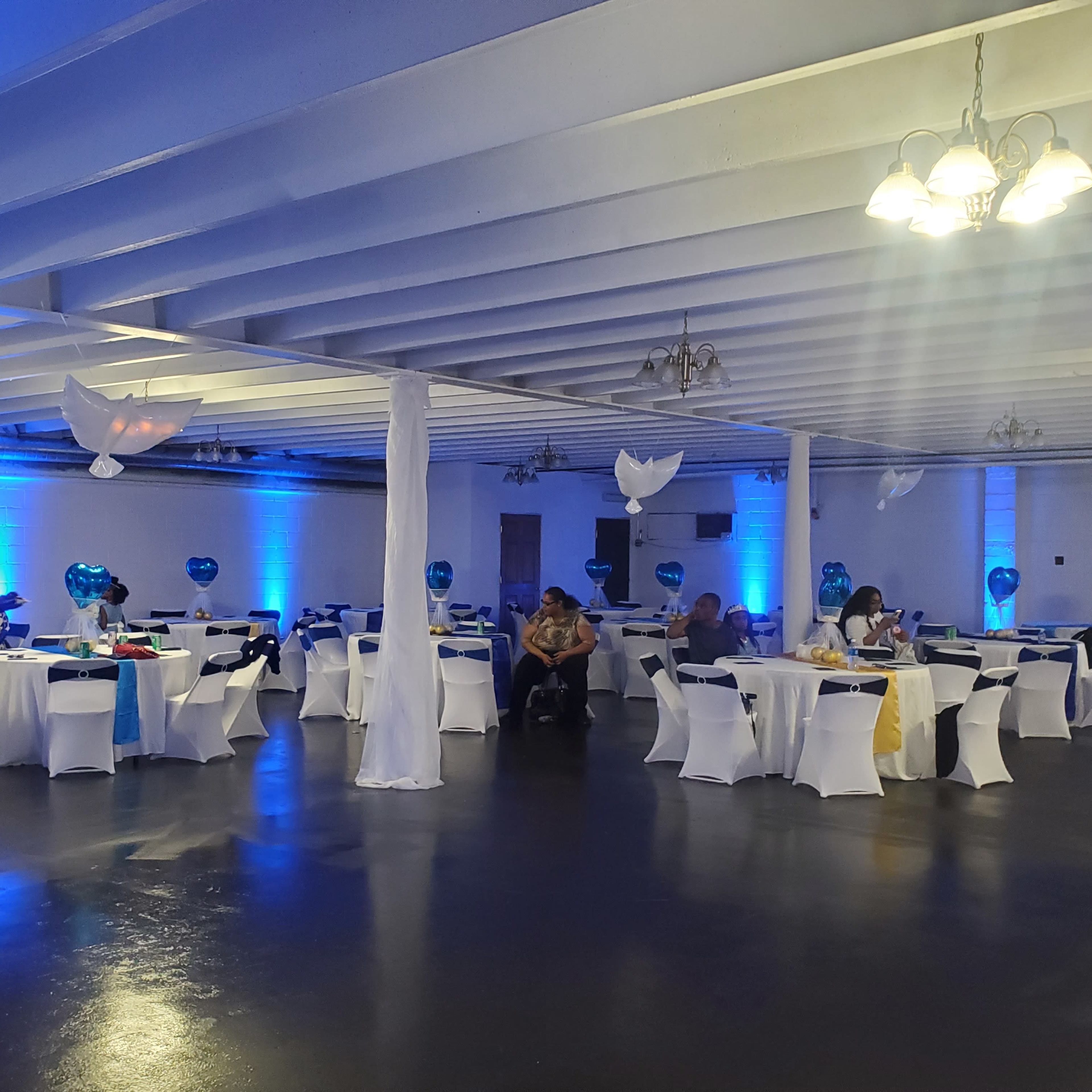 A banquet hall with decorated tables covered in white tablecloths and blue accents, illuminated by overhead lights and blue wall lighting.