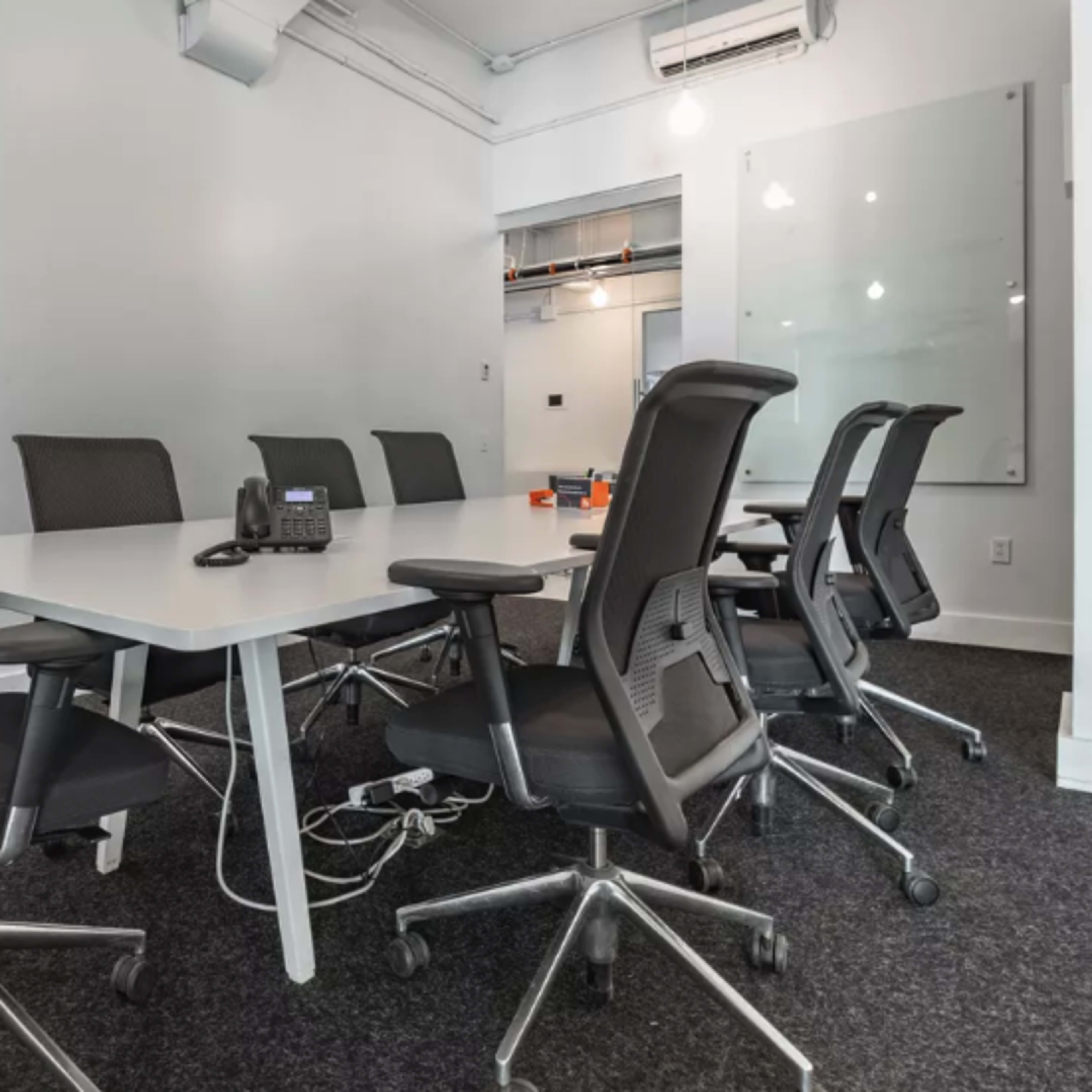 A modern conference room with a white table surrounded by six black office chairs and a phone on the table.