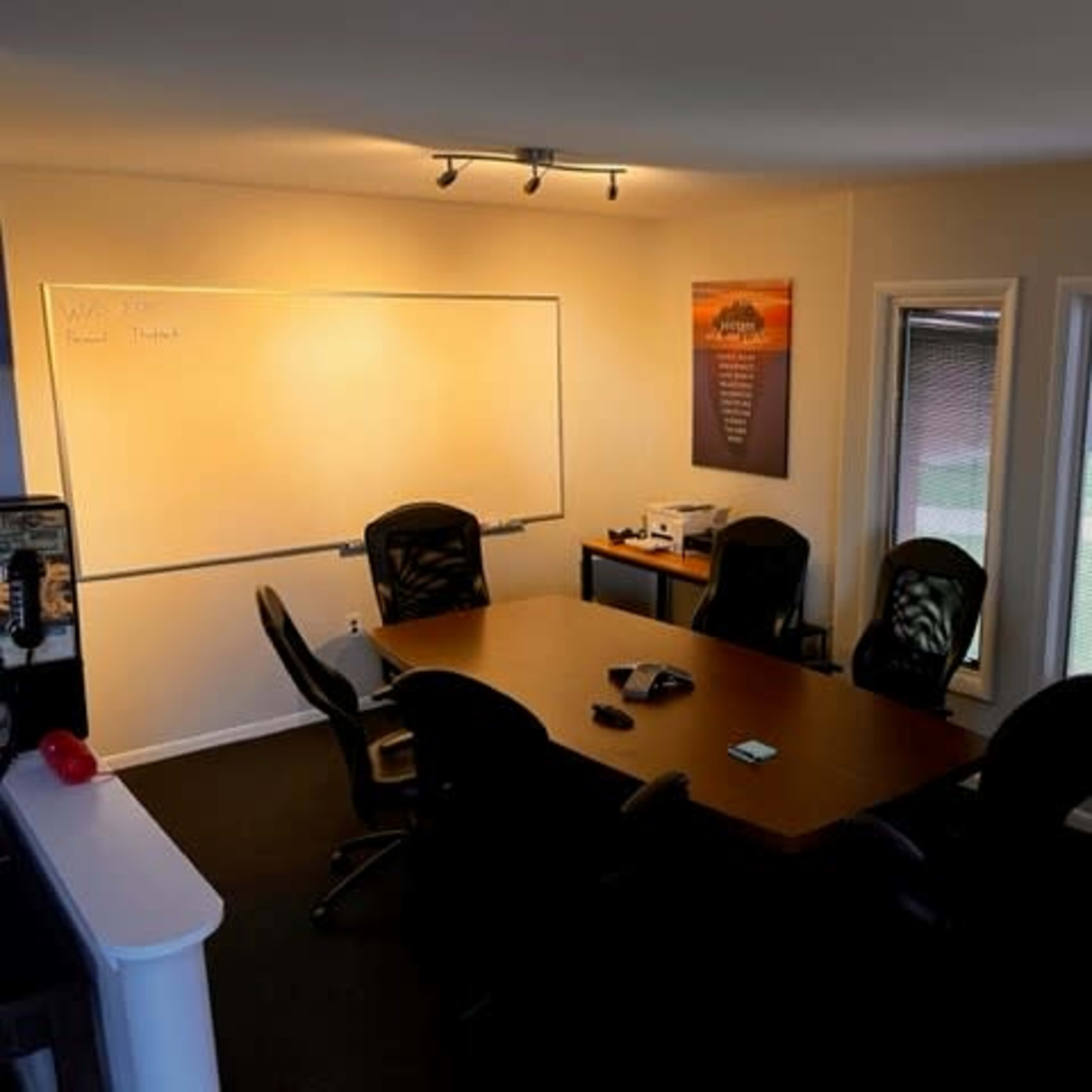 A meeting room with a large wooden table surrounded by black chairs, a whiteboard on the wall, a flip chart stand, and windows letting in natural light.