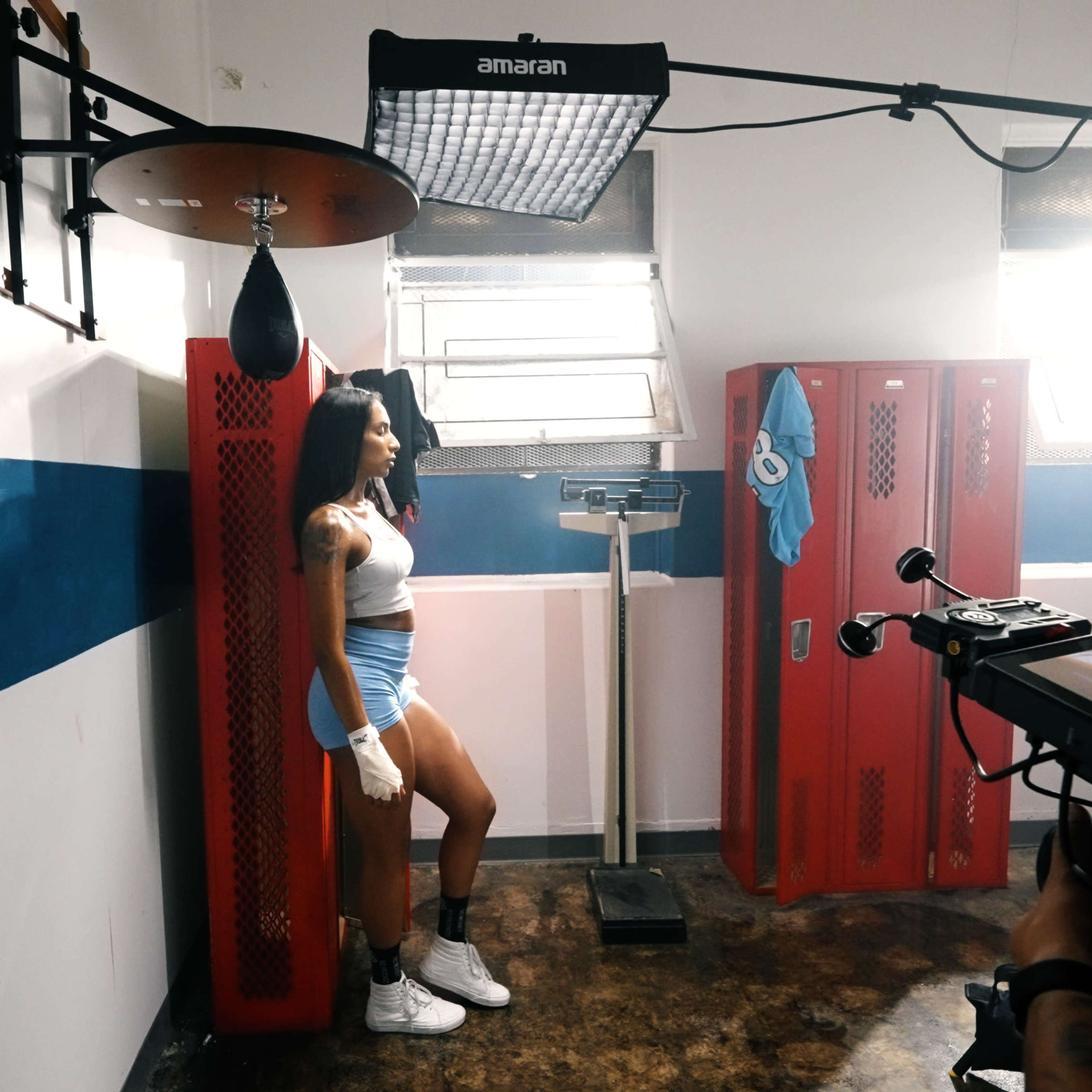 A person stands near a punching bag and scale in a dimly lit room with red lockers and a softbox light setup.