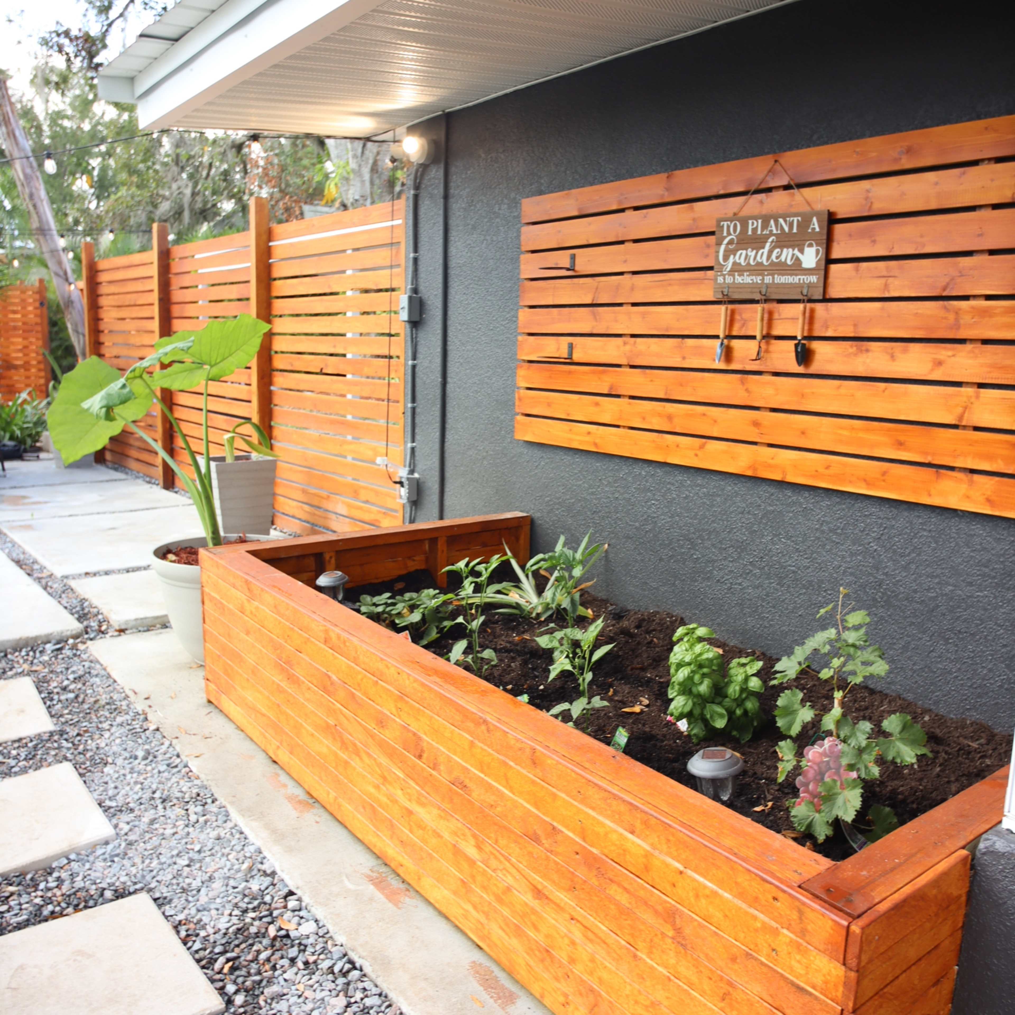 A wooden raised garden bed is positioned alongside a house, filled with soil and plants, with decorative fencing and stones leading to it.