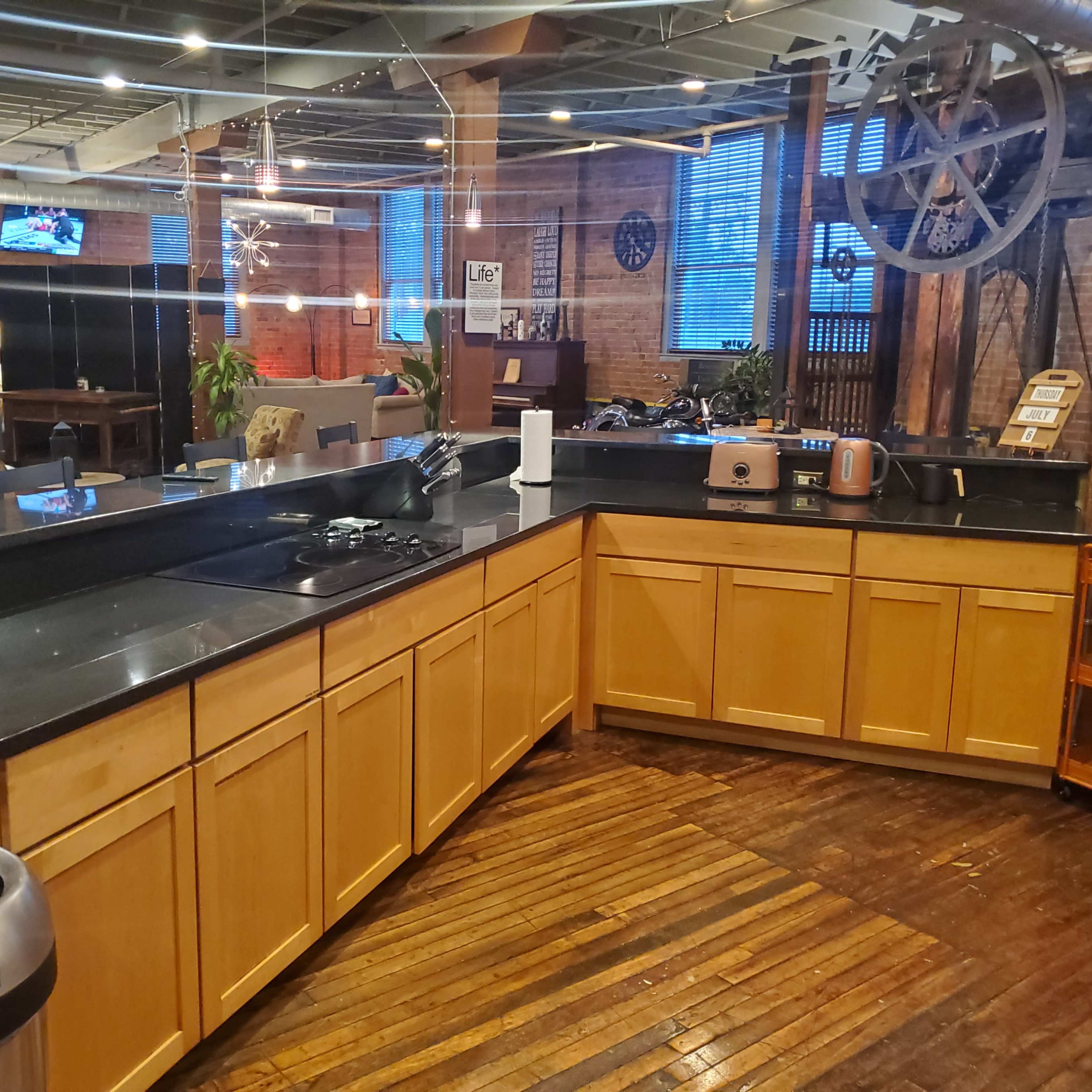 A modern kitchen with wooden cabinets, a sink, a stovetop, and various appliances, set in an industrial-style space featuring exposed brick walls and hardwood flooring.