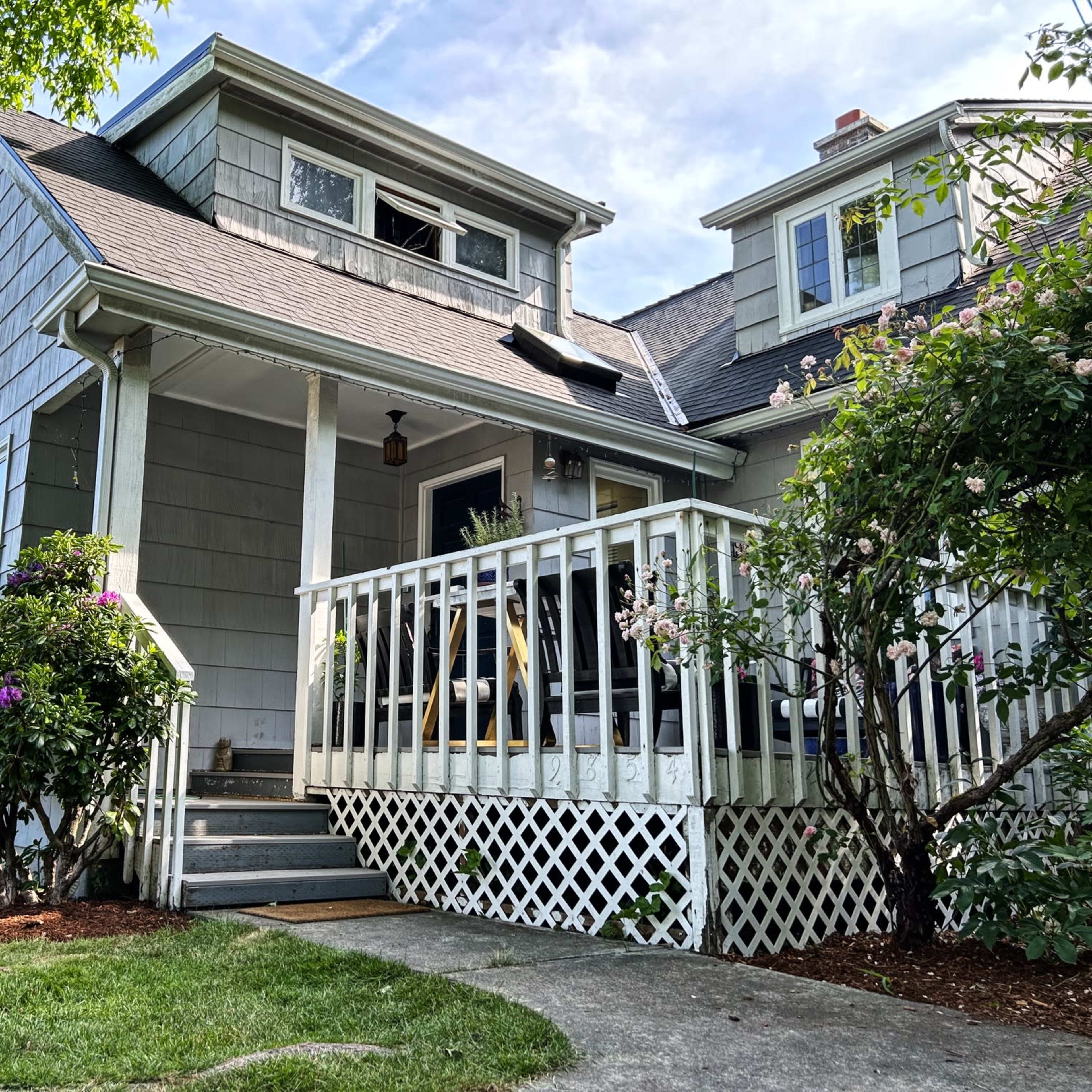 A two-story house with grey siding, a porch with a railing, and lush greenery surrounding the front yard.