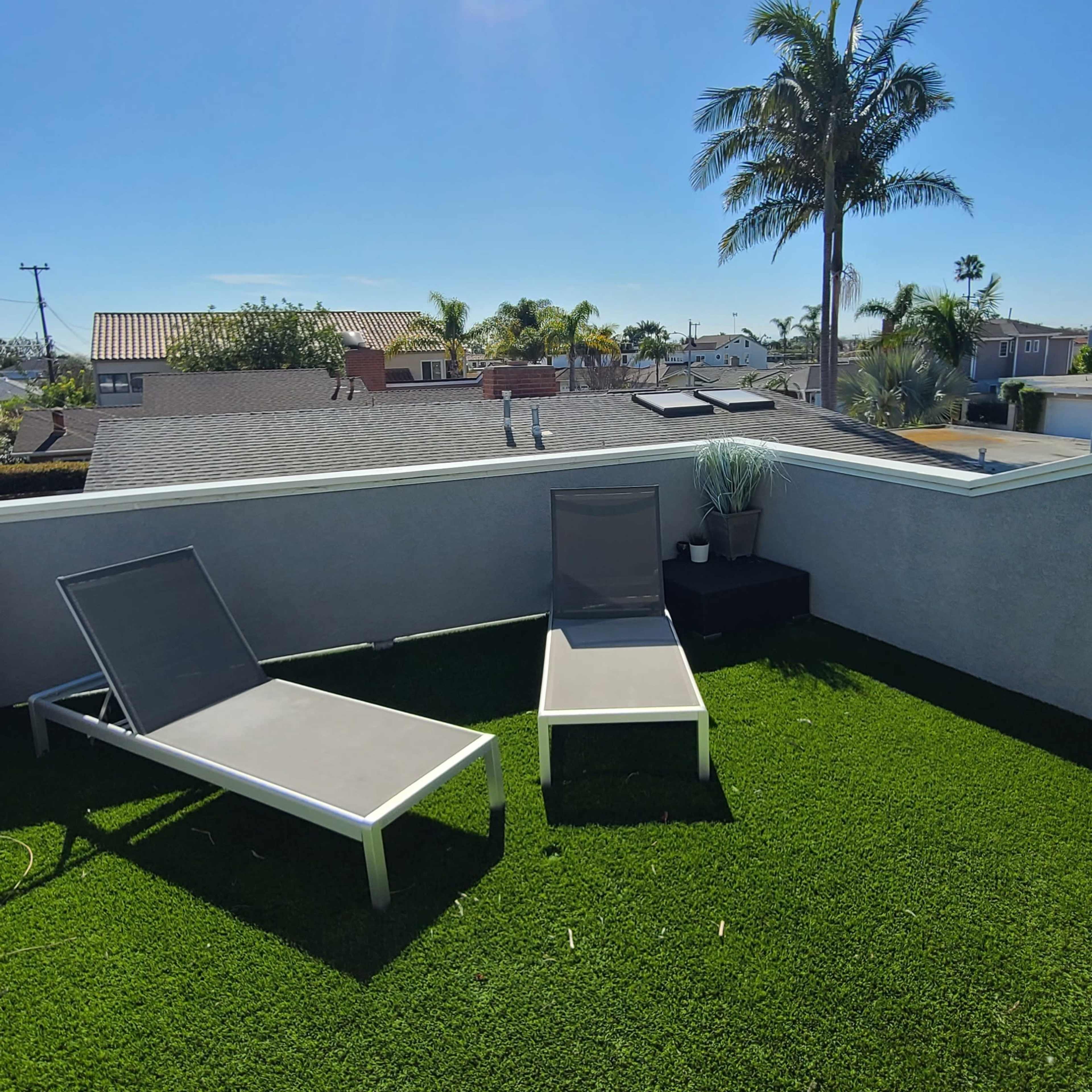 Two lounge chairs on a terrace with artificial grass, overlooking a neighborhood and palm trees under a clear blue sky.
