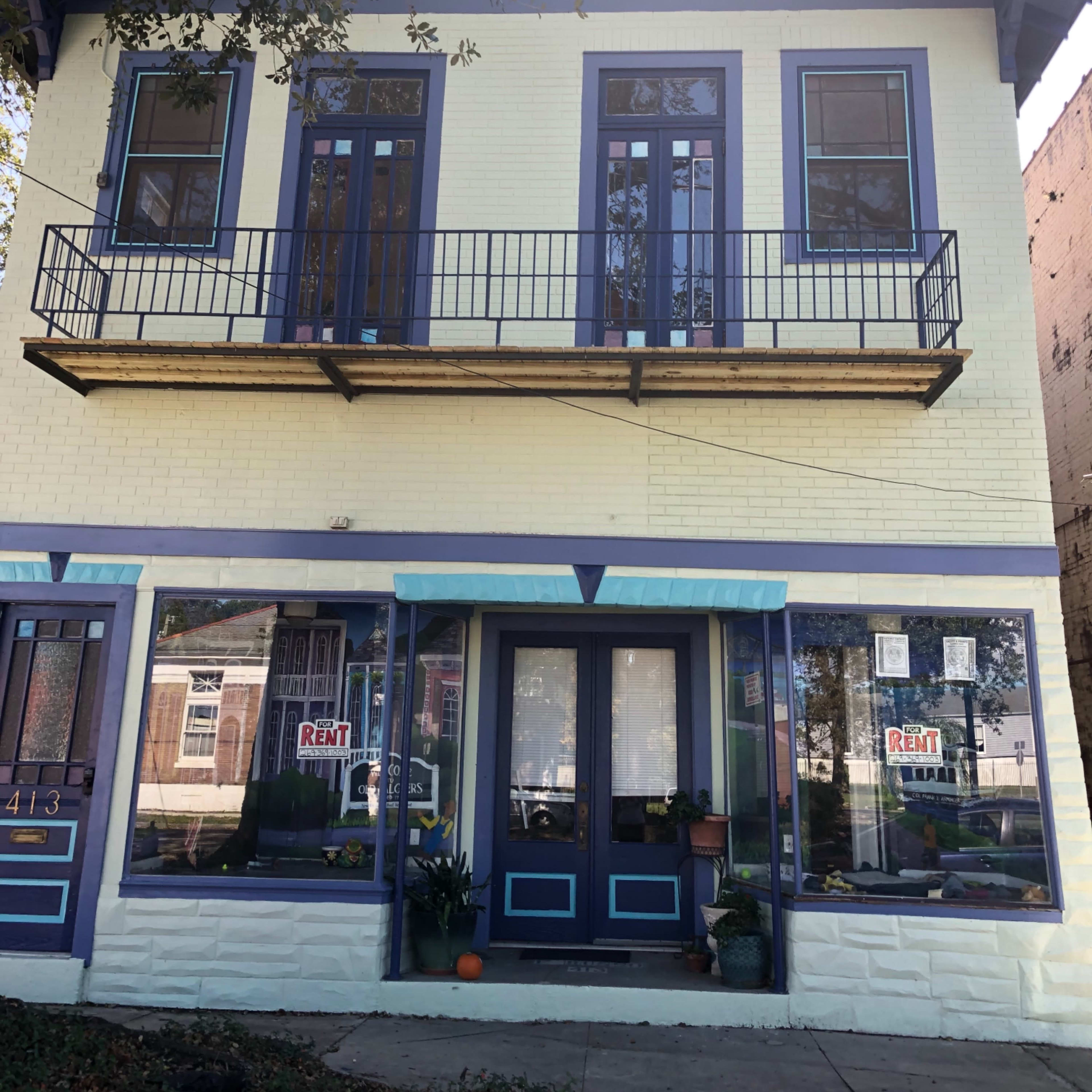 A two-story, yellow building with blue trim, featuring large windows and a "For Rent" sign in the front.