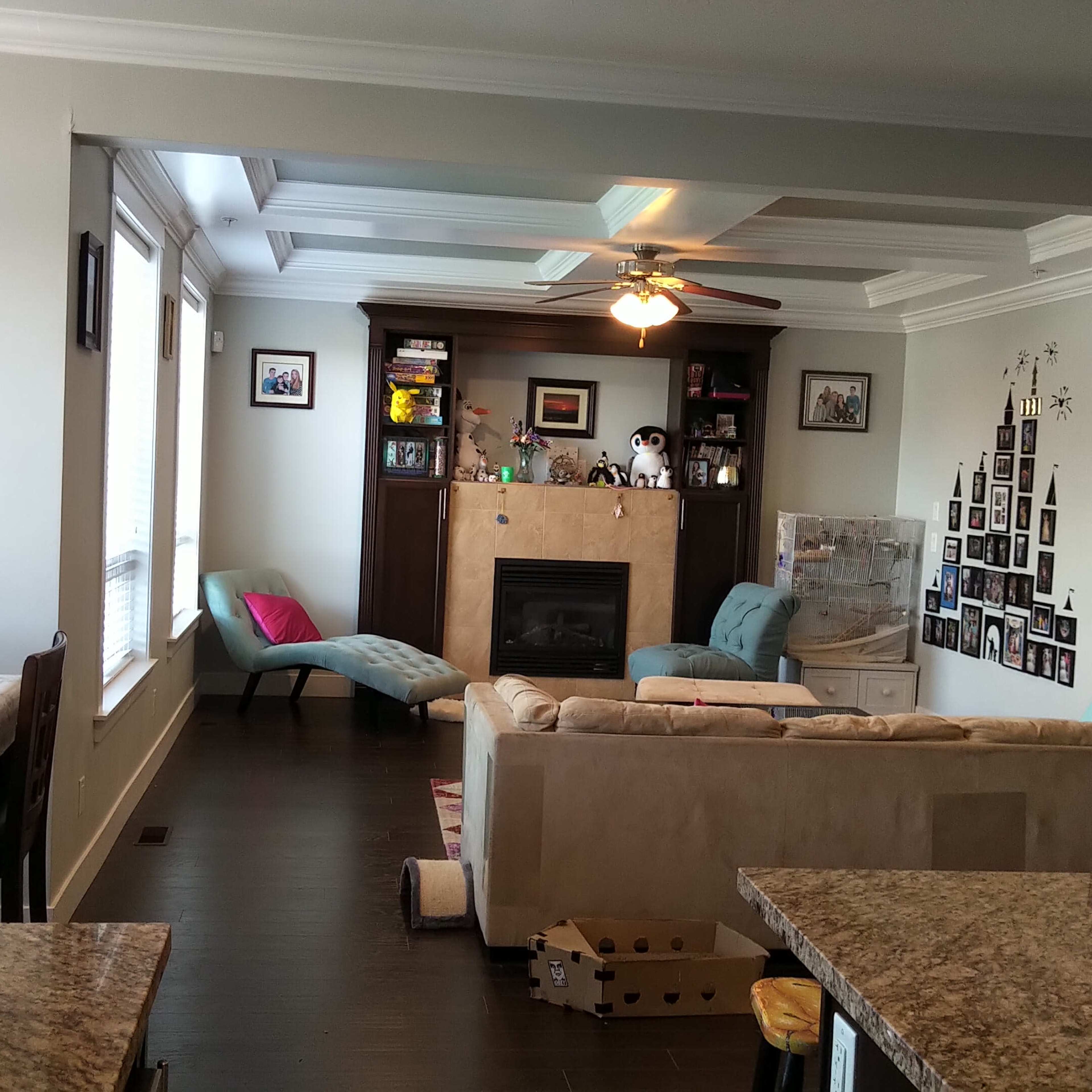 A modern living room with a cream-colored sofa, a fireplace, and various decorations on the walls, while a kitchen area with dark cabinets and a dining table is visible in the foreground.
