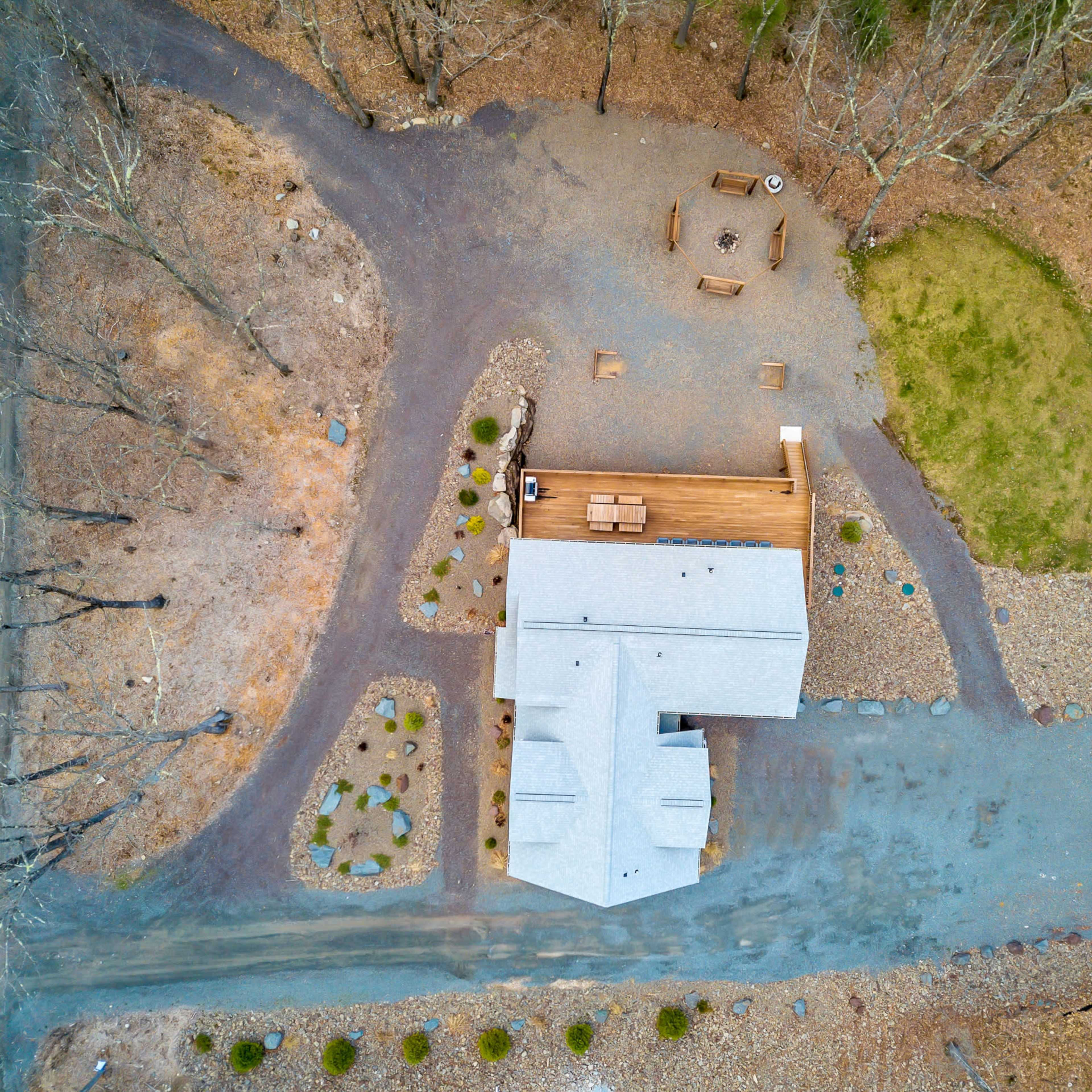 An aerial view of a house surrounded by a circular driveway, a fire pit area, and landscaped stones, with trees in the background.