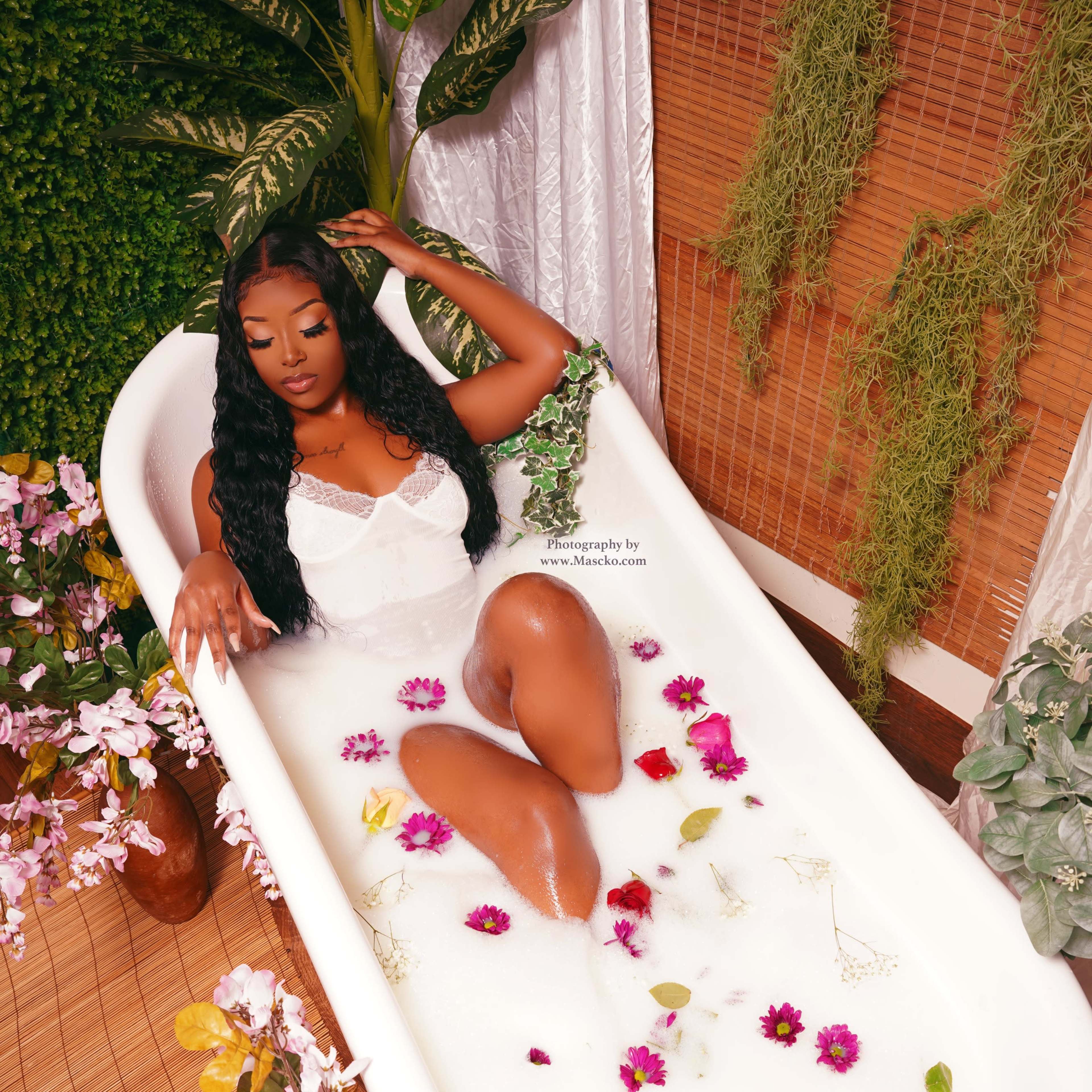 A person is relaxing in a milk bath filled with flowers and surrounded by plants in a beautifully decorated bathroom.