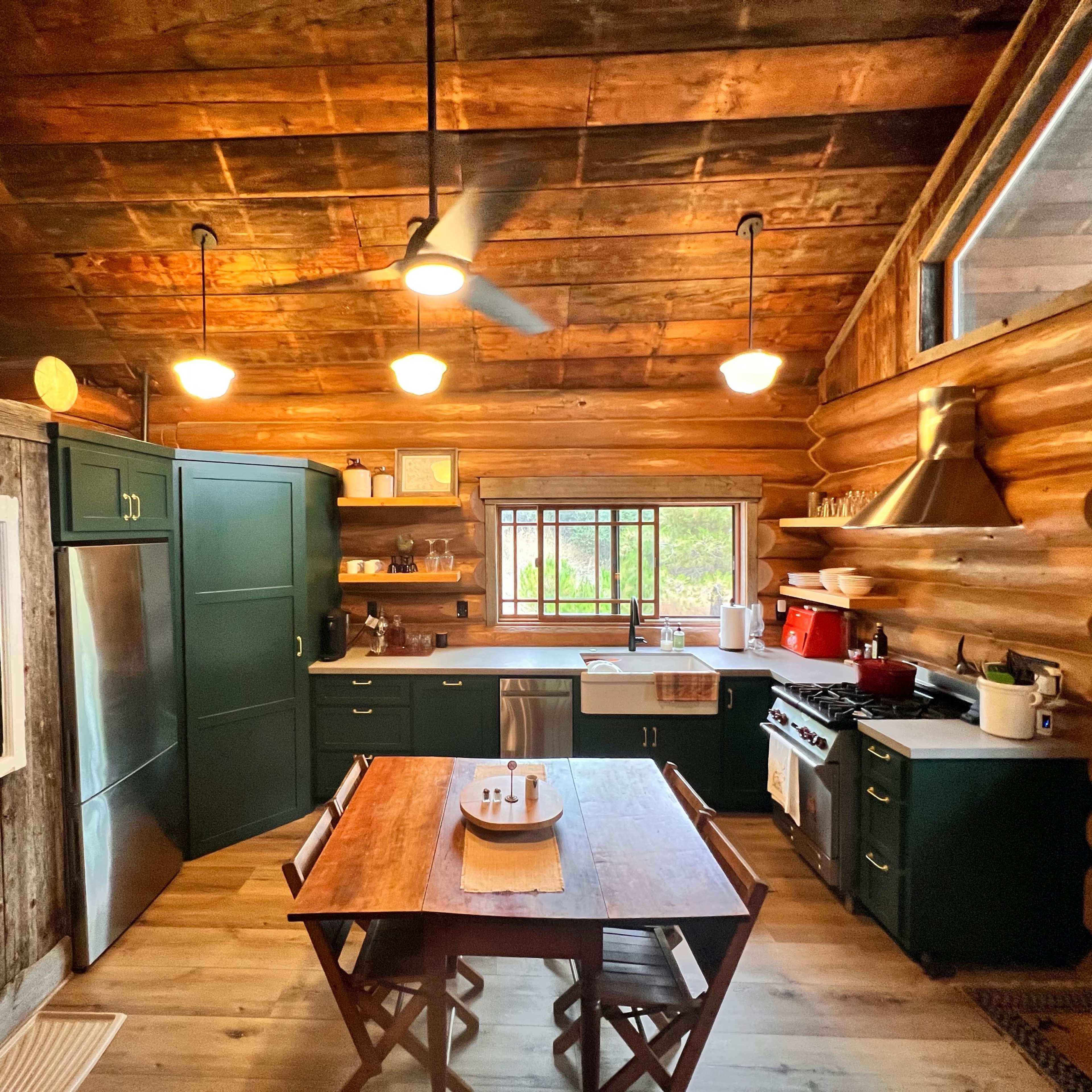 A cozy kitchen features a wooden log interior, green cabinetry, a dining table, and a large window providing natural light.