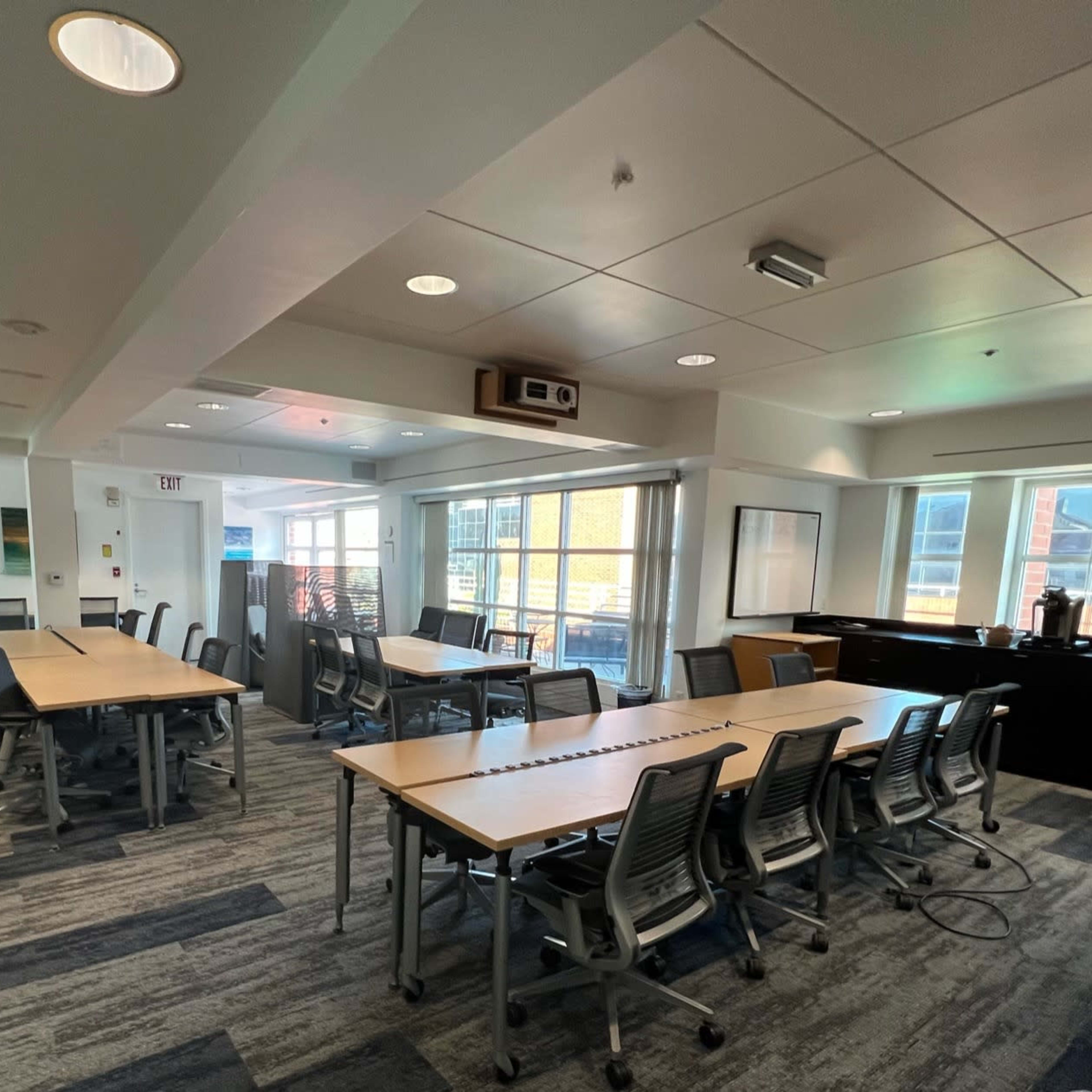 A modern conference room with multiple tables and chairs arranged around a central space, large windows, and a kitchenette area.