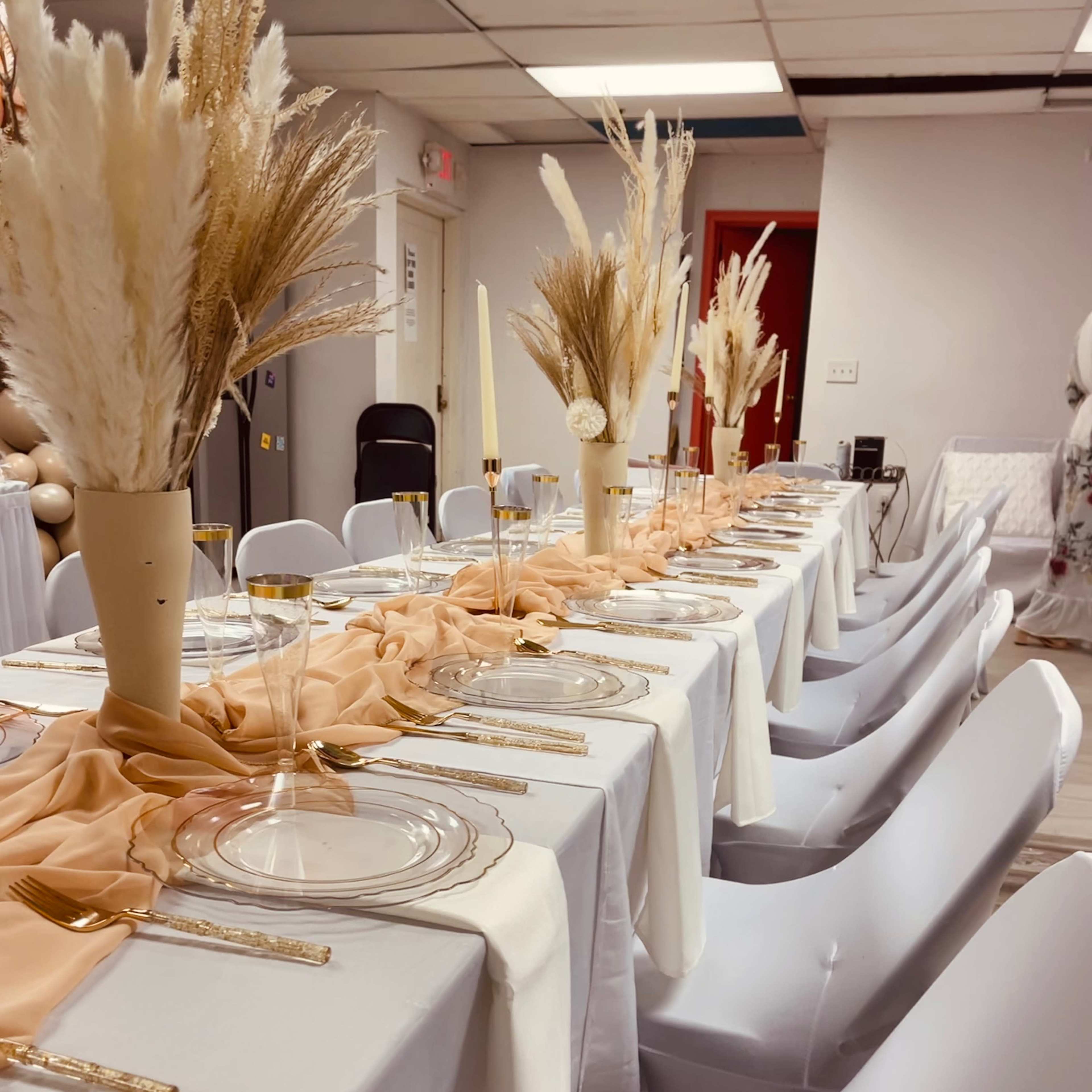 A long banquet table is set with white tablecloths, gold cutlery, and vases of dried flowers arranged in the center.